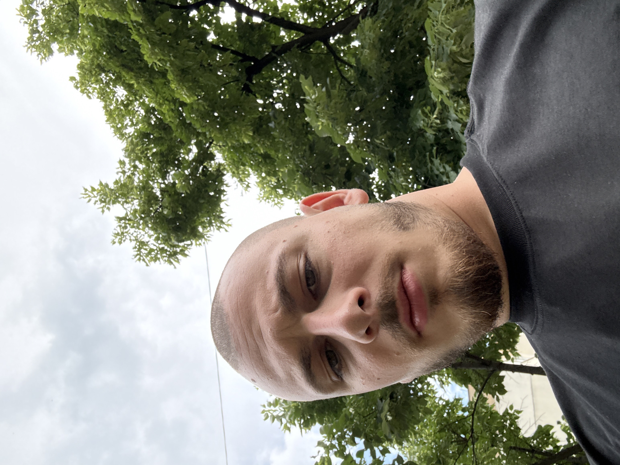 A low-angle, full-face selfie of a fair-skinned, bald or closely-shaved man with a beard, facing down at the camera. He is wearing a dark grey or black t-shirt. The background shows a cloudy sky and the green foliage of trees. The location cannot be definitively determined as Stara Zagora, Bulgaria, based on the image alone.