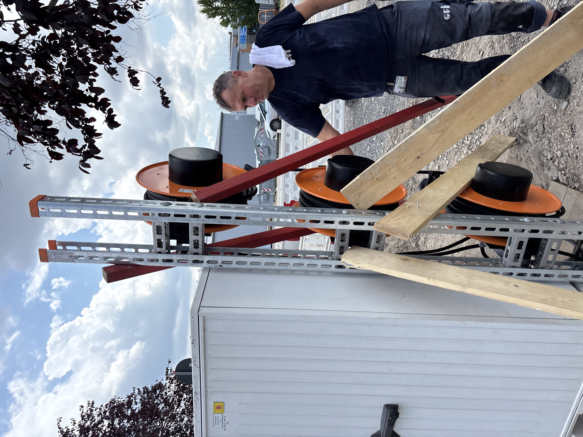 This is a medium shot, outdoor photograph of a man working on some equipment. The man, who appears to be middle-aged with short, graying hair, is wearing a dark blue t-shirt and dark pants. He has a white towel draped around his neck. He is leaning over some orange cable spools that are mounted on a silver metal rack. A red wooden plank is positioned diagonally across the spools, and another wooden plank is resting on top of the rack. The background shows a gravelly surface with some buildings and trees visible. The sky is cloudy with patches of blue. The lighting suggests it is daytime. There is some text visible on the man's pants which appears to be a logo or insignia. There is also a yellow sticker with text on the white container in the lower right of the frame. The location is identified as Mannheim, Germany. The overall impression is that of an industrial or construction site.