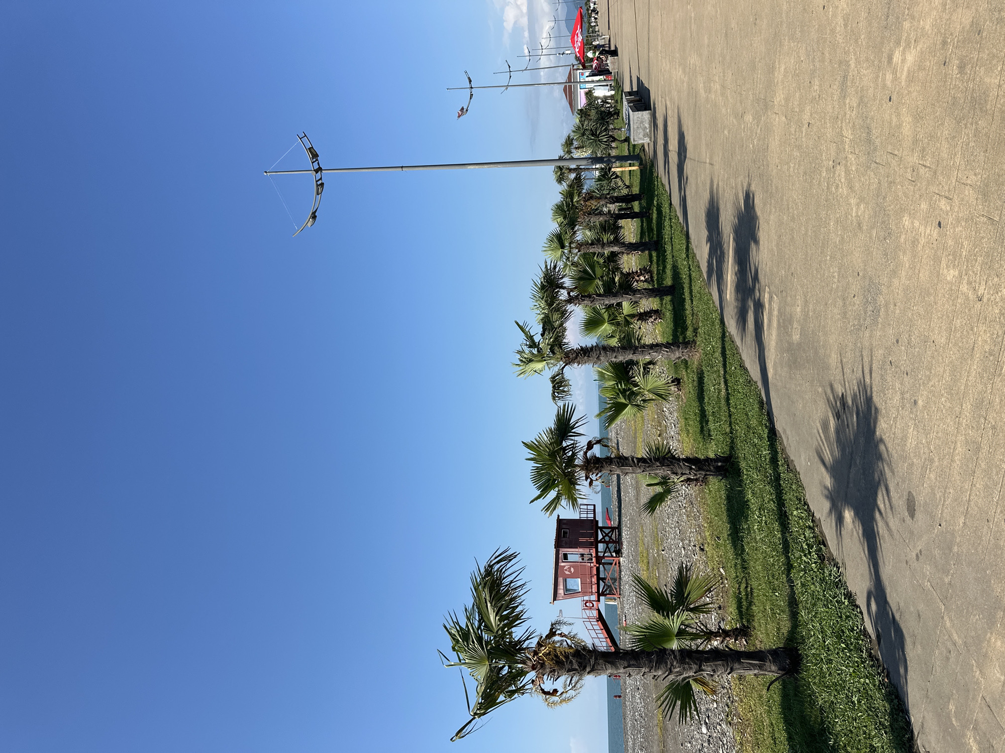The image depicts a seaside promenade in Batumi, Georgia, under a clear blue sky. The promenade runs parallel to a pebbly beach, separated by a strip of grass and a row of palm trees. On the beach, a red lifeguard station with stairs and a window is visible. Along the promenade, several tall lampposts line the path, casting shadows of palm trees on the concrete walkway. In the distance, there are a few people walking and a red umbrella over a vendor stall. The overall scene conveys a peaceful and sunny day at the coast.