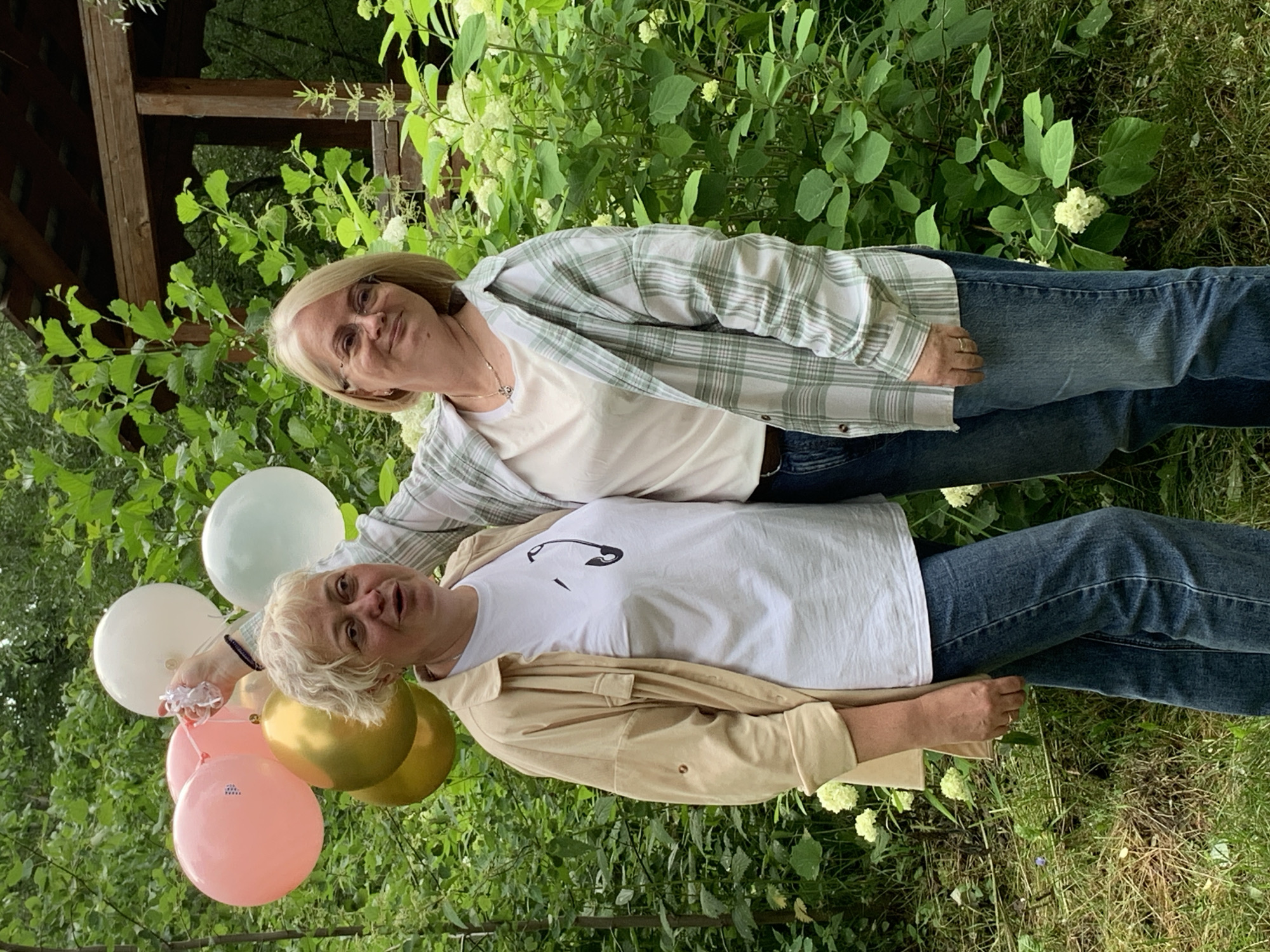 Two women are standing outdoors in what appears to be a garden or park setting. The woman in the foreground is wearing a tan jacket over a white t-shirt with a black design, and blue jeans. She is holding a bunch of balloons in various colors, including pink, gold, and white. The woman behind her is wearing a white t-shirt and a green and white plaid shirt, along with blue jeans. Both women have blond hair and appear to be smiling. The background is filled with lush green foliage and some white flowers. The lighting suggests it is daytime, likely a clear or partly cloudy day. The scene evokes a celebratory or festive mood, possibly a birthday or other special occasion. No visible text is present in the image.