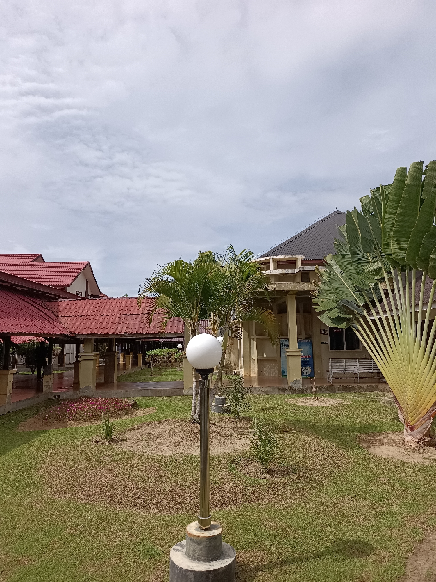A paved walkway with red tiled roofs and beige supporting pillars is visible to the left of the image, with a person walking away from the camera down the walkway. In the foreground, a lamp post with a spherical light fixture stands in a grassy area. Behind the walkway, a building with a dark grey roof and beige walls is visible. A large traveler's palm tree with fan-shaped leaves is situated to the right of the building. The sky above is cloudy. The location is the city of Mesjid Reuntoh, Indonesia.