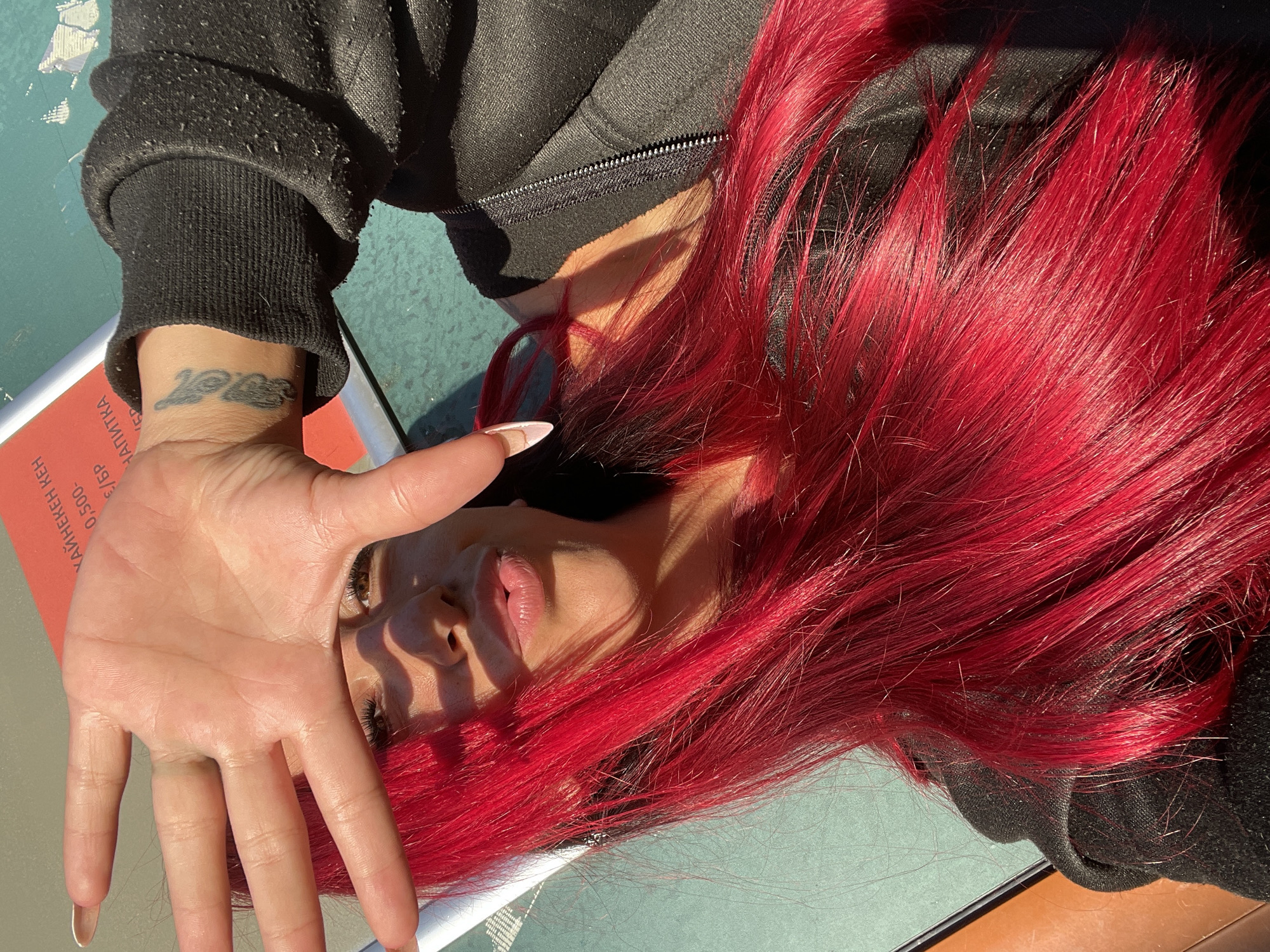 A young woman with vibrant red hair covers her face with her left hand, revealing her eyes, nose, and lips as she angles her head towards the camera. She is wearing a black zip-up jacket. A tattoo is visible on her right wrist. The background consists of a green surface and a red object with some text in Cyrillic script, suggesting she is likely outdoors in a public space, possibly in Sofia, Bulgaria, based on the Cyrillic writing. The lighting indicates it is daytime.