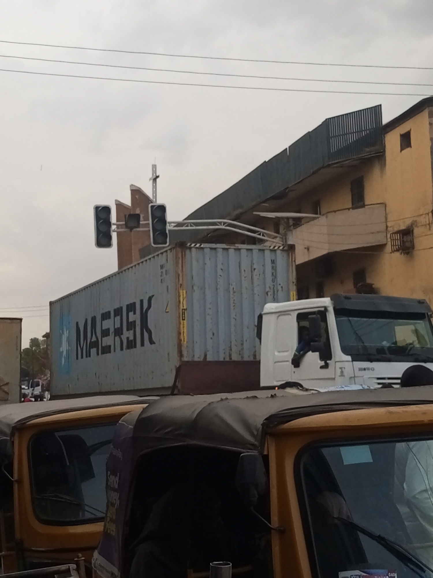 The image depicts a street scene in Maiburiji, Nigeria, with a Maersk shipping container on a white truck. Traffic lights are visible overhead. In the foreground, there are several auto rickshaws, also known as "keke" in Nigeria. A multi-story building is in the background, next to the traffic light. The sky is overcast.