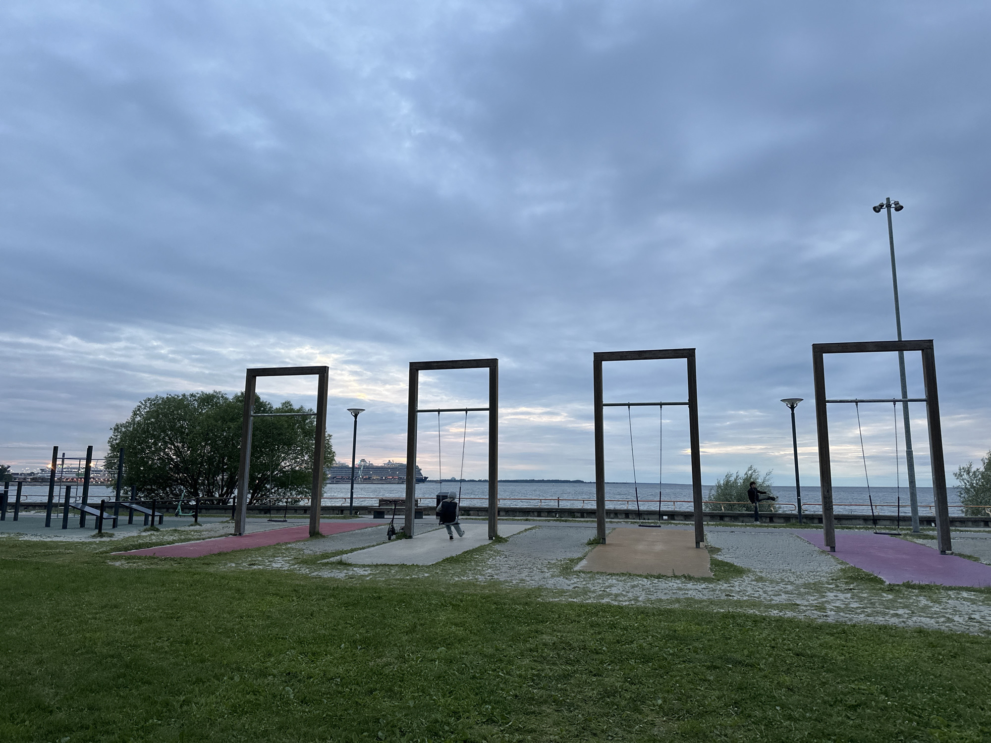 The image captures a waterfront playground in Tallinn, Estonia, overlooking the Baltic Sea. The sky is overcast with a muted blue and grey hue. In the foreground, a lush green lawn transitions to a gravel area with four distinct swing sets, each featuring a wooden frame. The swing sets are spaced evenly along the waterfront.
One swing set has a child actively swinging, their back to the camera, dressed in dark clothing and a grey hat. Another swing set is occupied by a person standing near it, facing the sea, wearing dark clothing. The remaining two swing sets are empty.
Beyond the playground, the sea stretches to the horizon, with a large cruise ship visible in the distance. The ship is white with red accents and appears to be docked or moving slowly. The coastline is lined with a low wall and some vegetation.
To the left of the swing sets, a section of an outdoor gym equipment area is visible, including parallel bars and other fitness apparatus. A lone green scooter rests on the gravel near the gym equipment. The overall atmosphere is calm and serene, suggesting it is either early morning or late evening. The playground appears well-maintained and situated in a public park area.