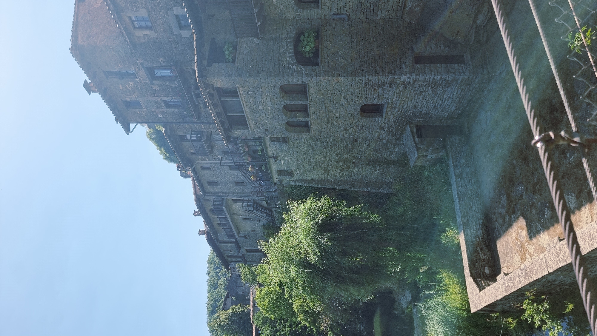 This is a vertical, eye-level, exterior shot of a stone building in Rupit i Pruit, Spain. The building is constructed of rough-hewn grey stone blocks with a terracotta tile roof. It has multiple levels with balconies, arched windows, and small balconies with wrought-iron railings. Green foliage, including trees and ivy, grows around and on the building. In the foreground, a stone wall encloses a courtyard area that is partially in shadow. A cable railing runs across the bottom of the image. The sky is clear and blue.
