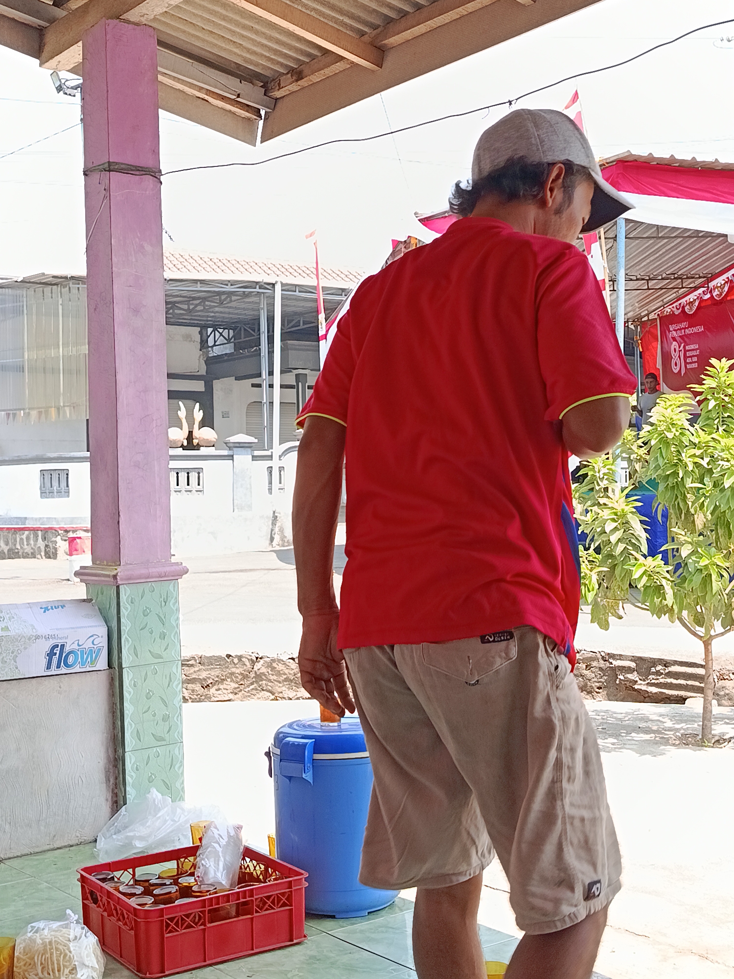 A man wearing a red t-shirt and tan shorts stands facing away from the camera, his head turned slightly to the side. He is wearing a gray baseball cap. The man is positioned near a blue cooler and a red plastic crate filled with small containers and plastic bags of what appear to be food items. Behind him, a white building with decorative statues on top can be seen, along with some vegetation. The scene appears to be outdoors, with a covered structure overhead supported by a pink pillar. There are Indonesian flags visible in the background, suggesting a local event or celebration. The environment looks like an outdoor market or food stall area in Jurug, Indonesia, a district known for its batik and tourism.
