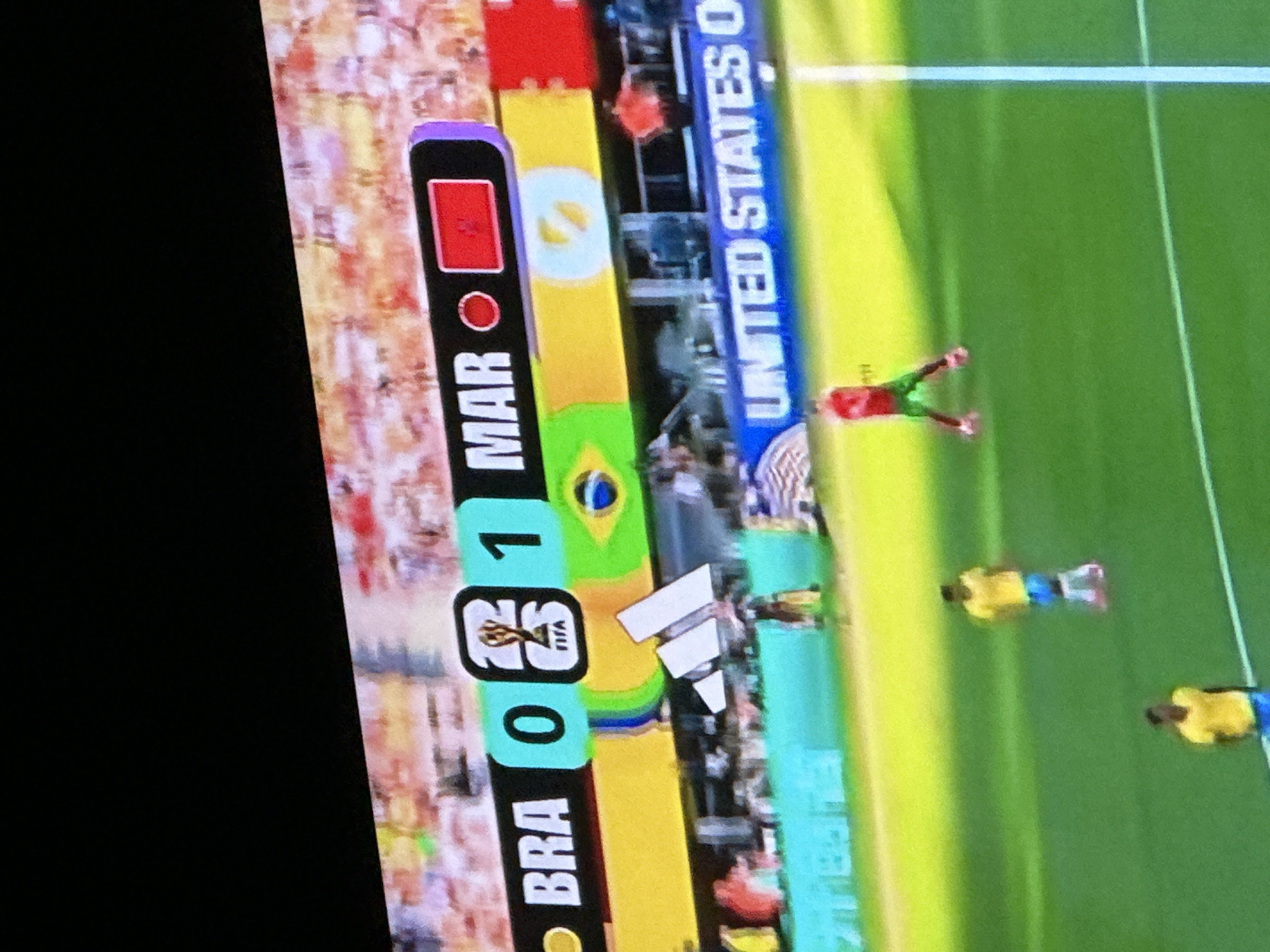 The image shows a view of a soccer match being displayed on a screen, likely a television or projector. The screen shows a scoreboard with "BRA 0" and "MAR 1" displayed. The "MAR" side also has a red flag icon. Below the scoreboard, there are graphics of national flags, including the Brazilian flag, and the Adidas logo. Several blurred figures of soccer players in uniform are visible on a green field. The upper portion of the image shows a dark area, possibly the edge of the screen or a darkened room. The scene suggests a broadcast of a soccer game, with the score indicating Morocco is leading Brazil 1-0. The image does not contain any information or visual cues that would allow for the identification of the city of Rayfield, Nigeria.