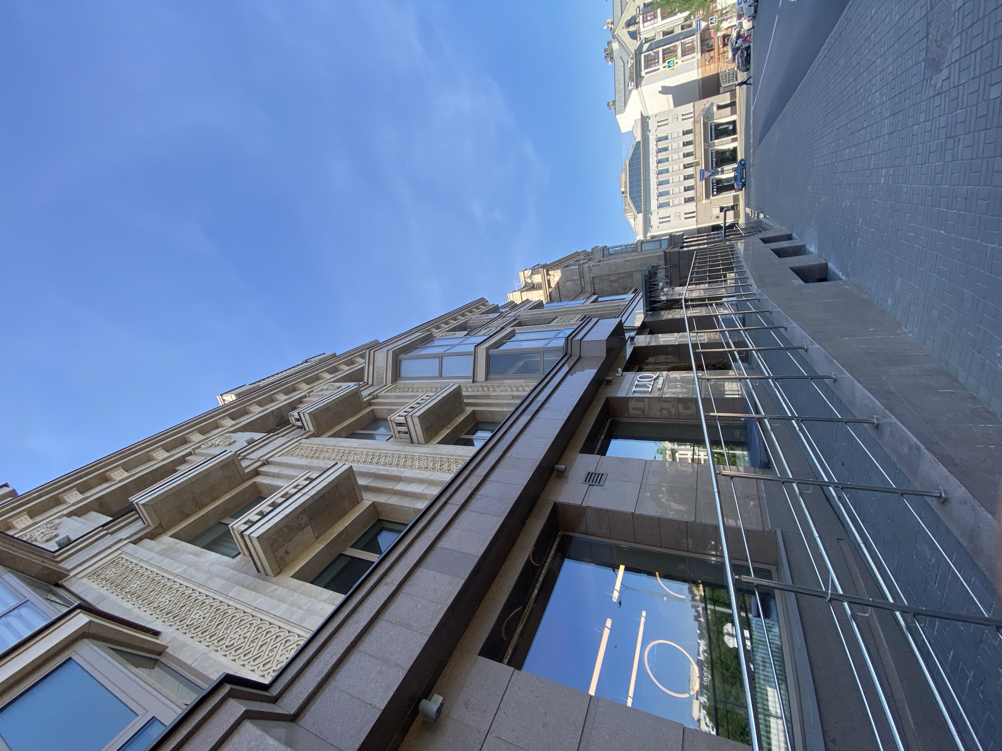 The image shows an upward-angled view of a building in Kazan, Russia, under a clear blue sky. The building is multi-storied, constructed with light-colored stone and ornate detailing. It features large windows with reflective surfaces, and protruding architectural elements such as balconies or decorative ledges. A portion of another building and a street are visible in the upper right corner of the image, suggesting a location in an urban area. The street is paved with grey brick pavers. The architecture appears modern with classical influences.