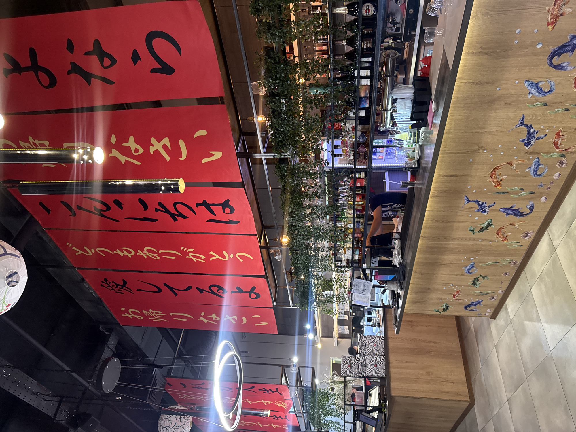 The image shows the interior of a Japanese restaurant. Above, there are red banners with Japanese writing. Below, there are hanging circular light fixtures. In the background, a bar is visible with bottles of alcohol. In the distance, there are patrons sitting at the bar. The location is Domodedovo, Russia.