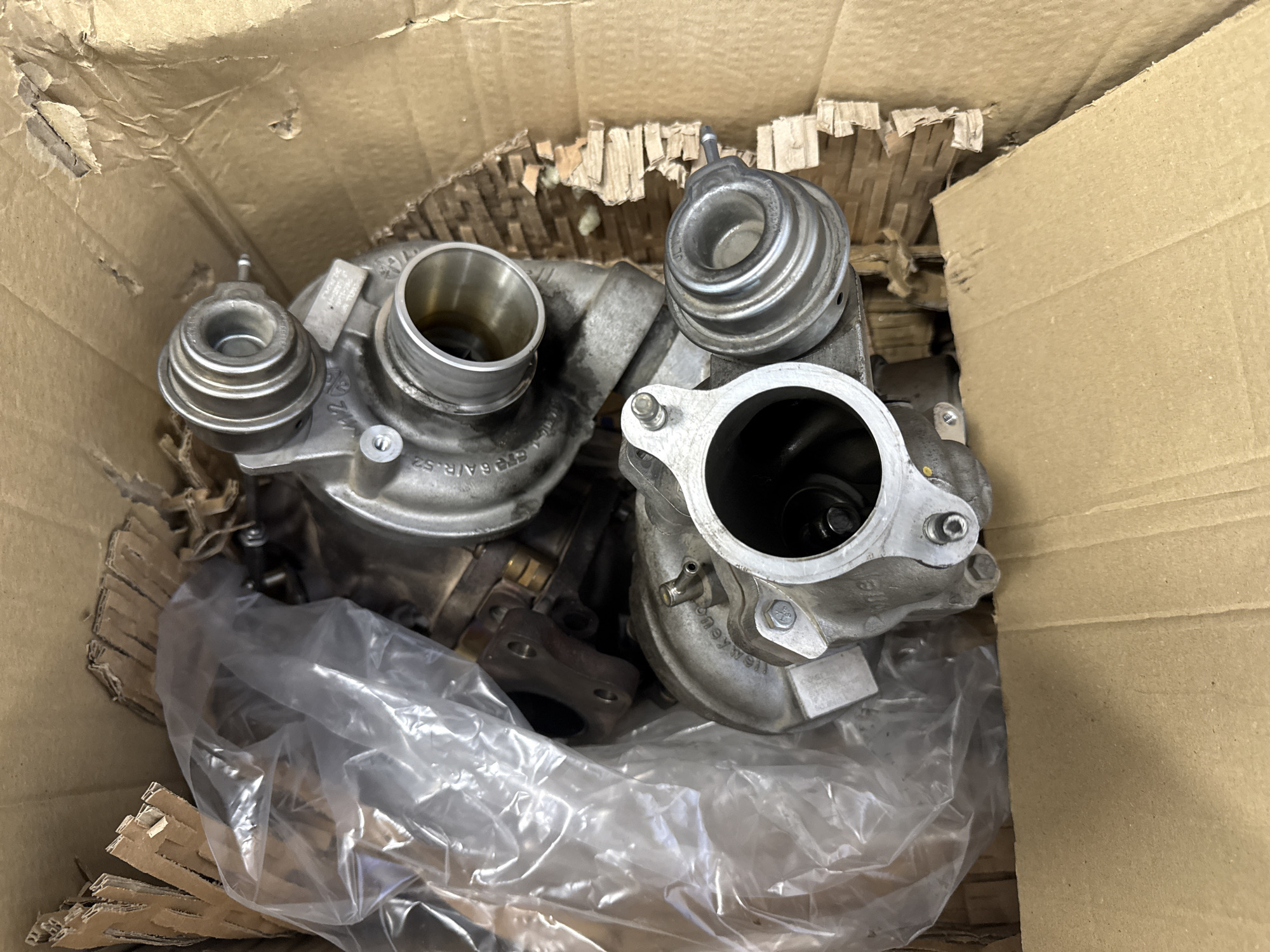 The image shows two turbochargers in a cardboard box. The box contains some packing material, including shredded cardboard and a clear plastic bag. The turbochargers are gray and metallic. One has a circular opening facing the viewer, revealing the turbine blades inside. The location is unknown, so there is no mention of Varna, Bulgaria.
