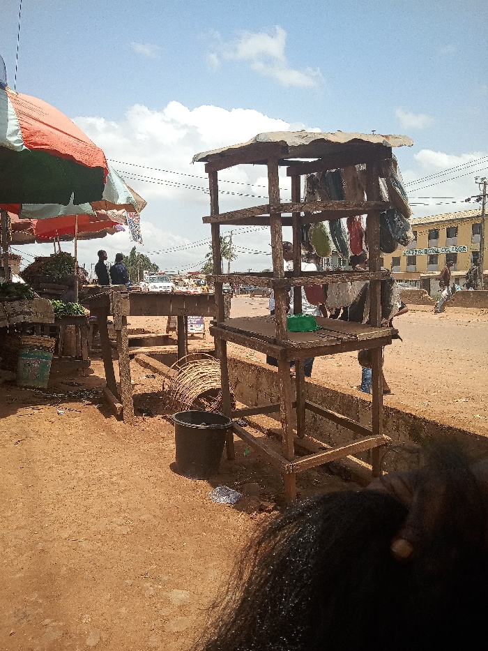 The image depicts a street scene in Ota, Nigeria, located at coordinates 6.683 latitude, 3.177 longitude. A large, multi-colored striped umbrella covers a makeshift stall with produce, while a wooden stand with goods hanging from it is located to the right of the stall. A person can be seen walking away from the wooden stand and another person is standing near it, out of focus. Several other people are standing or walking in the background. The ground is dirt, and there is a black plastic bucket, a roll of wire and a few other objects visible in the foreground. The scene is framed from a low angle, and a long strand of black hair is visible in the foreground. The background shows a building with "Adeyemi Royal College" sign on the front, as well as a road and other buildings. The scene is likely an outdoor market or street vendor area.