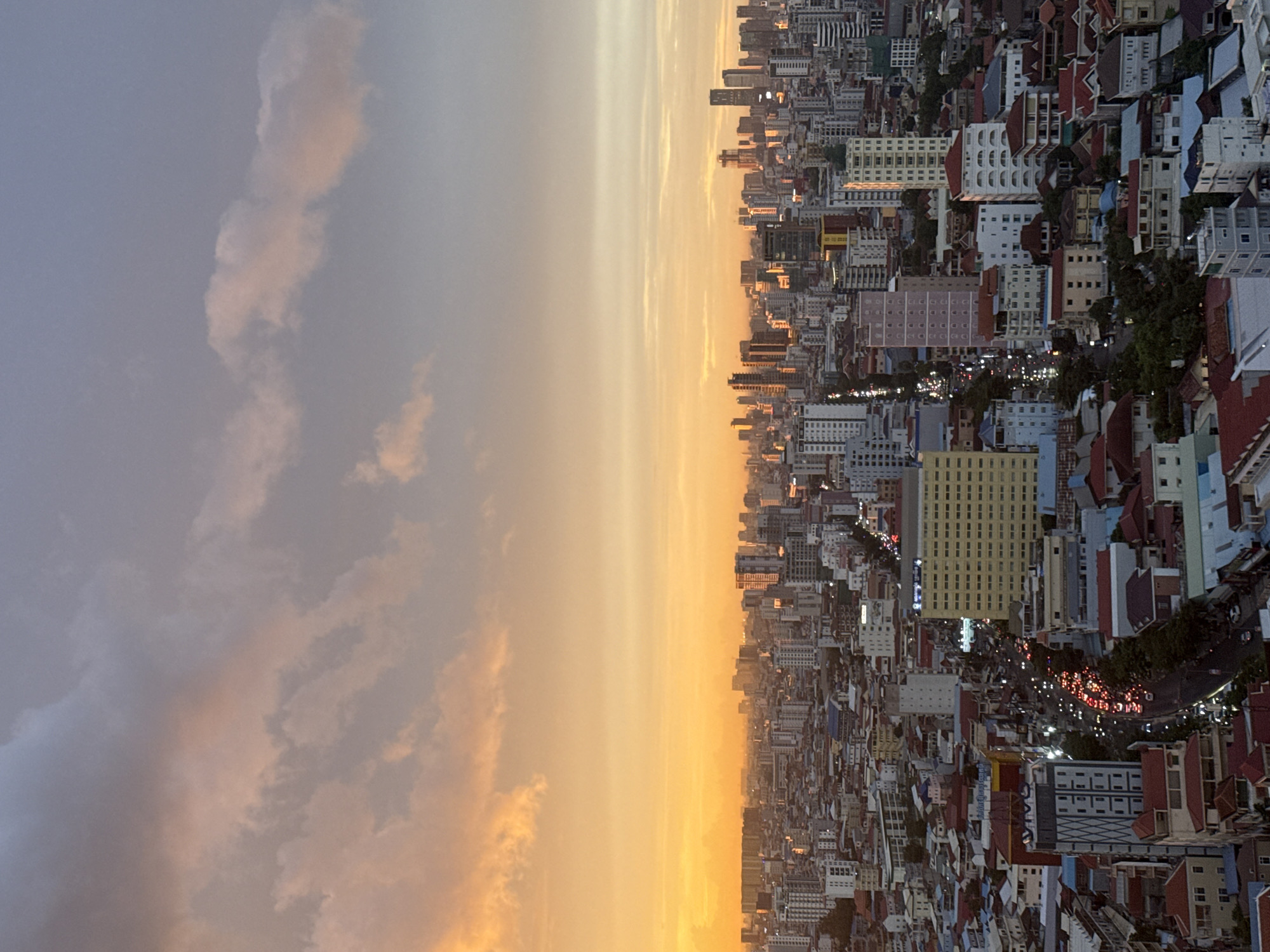 The image is a high-angle view of a densely populated urban landscape at sunset, likely in Khan Mean Chey, Cambodia, given the provided context. The sky dominates the upper half of the frame with a gradient of warm oranges and yellows near the horizon, transitioning into soft pinks and muted grays higher up, indicative of sunset lighting. Wispy clouds are scattered across the sky, catching the fading sunlight.
Below the sky is a vast expanse of city buildings, characterized by a mix of modern high-rises and lower-density residential structures. The architecture appears to be a blend of concrete and glass, with many buildings featuring flat roofs. The sheer density of the buildings suggests a significant urban center.
A prominent feature in the middle ground is a wide, busy street, illuminated by the headlights and taillights of numerous vehicles. The red glow of taillights indicates traffic flow, creating streaks of light down the road. The surrounding buildings along this street vary in height and architectural style, with some exhibiting colorful facades and others appearing more utilitarian. One notable building is a large, rectangular structure with a pale yellow facade, potentially an office building or commercial center, situated on the left side of the busy street.
The lower portion of the image reveals more residential areas with smaller buildings, some featuring red-tiled roofs. Trees and greenery are visible, interspersed between the buildings, suggesting that this is not a purely concrete jungle. The overall impression is of a vibrant, active city at dusk, with the warm hues of the sunset casting a serene glow over the bustling urban environment. No individuals are clearly discernible in the image due to the distance and the low light conditions.
