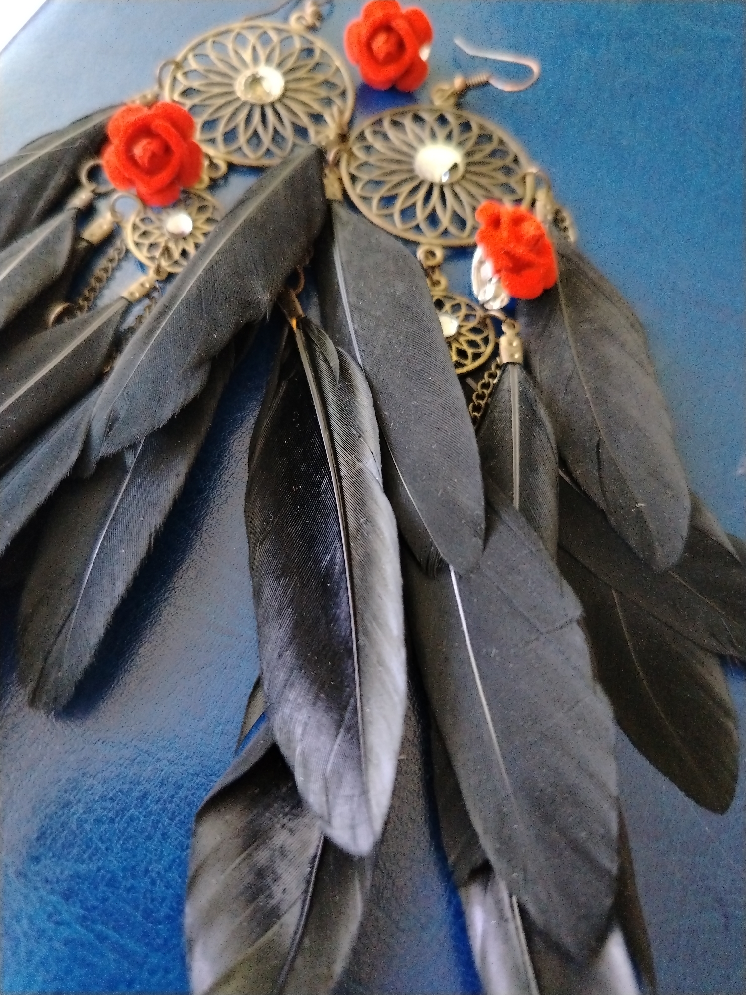 The image displays a close-up of an ornate earring featuring black feathers, red floral accents, and metallic filigree with clear rhinestones. The earring is positioned on a textured blue surface, likely a fabric or leather material. The composition is tight, focusing on the intricate details of the jewelry. There are no people, actions, or interactions depicted. The image does not contain any information that would allow for the identification of its location in Krasnodar, Russia.