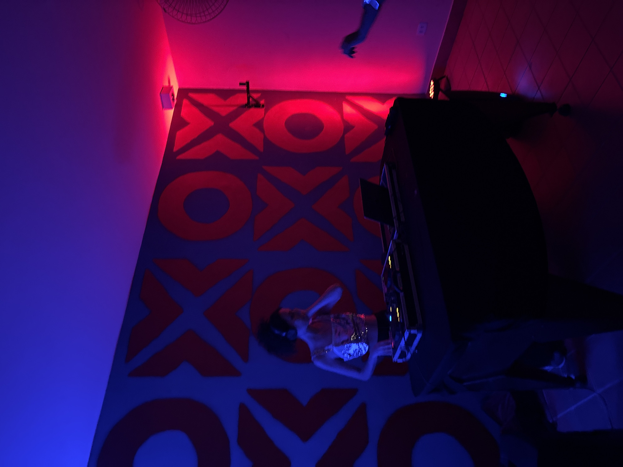 The interior is a nightlife venue in São Paulo, Brazil, featuring an XOXO pattern floor and vibrant red and blue lighting. A DJ is performing, positioned at a console with equipment. There is another arm raised in the background.