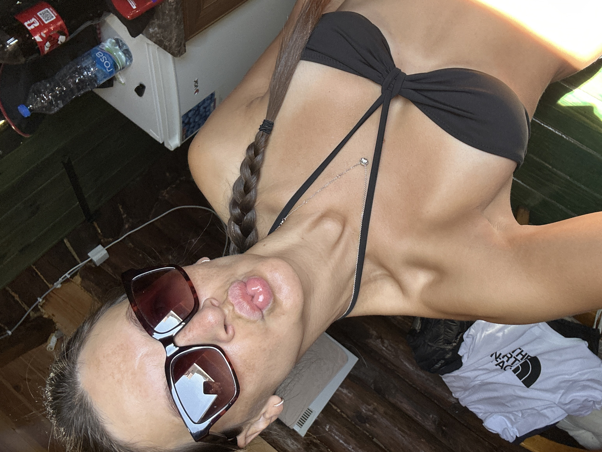 An outdoor selfie shows a woman posing with her lips puckered, wearing a black bikini top, sunglasses, and a delicate necklace. Her hair is braided and falls over her shoulder. In the background, a wooden structure is visible, along with a small white refrigerator and a plastic bottle of water. A white t-shirt with a black logo is also present in the scene. The overall impression is of a casual, sun-drenched setting.