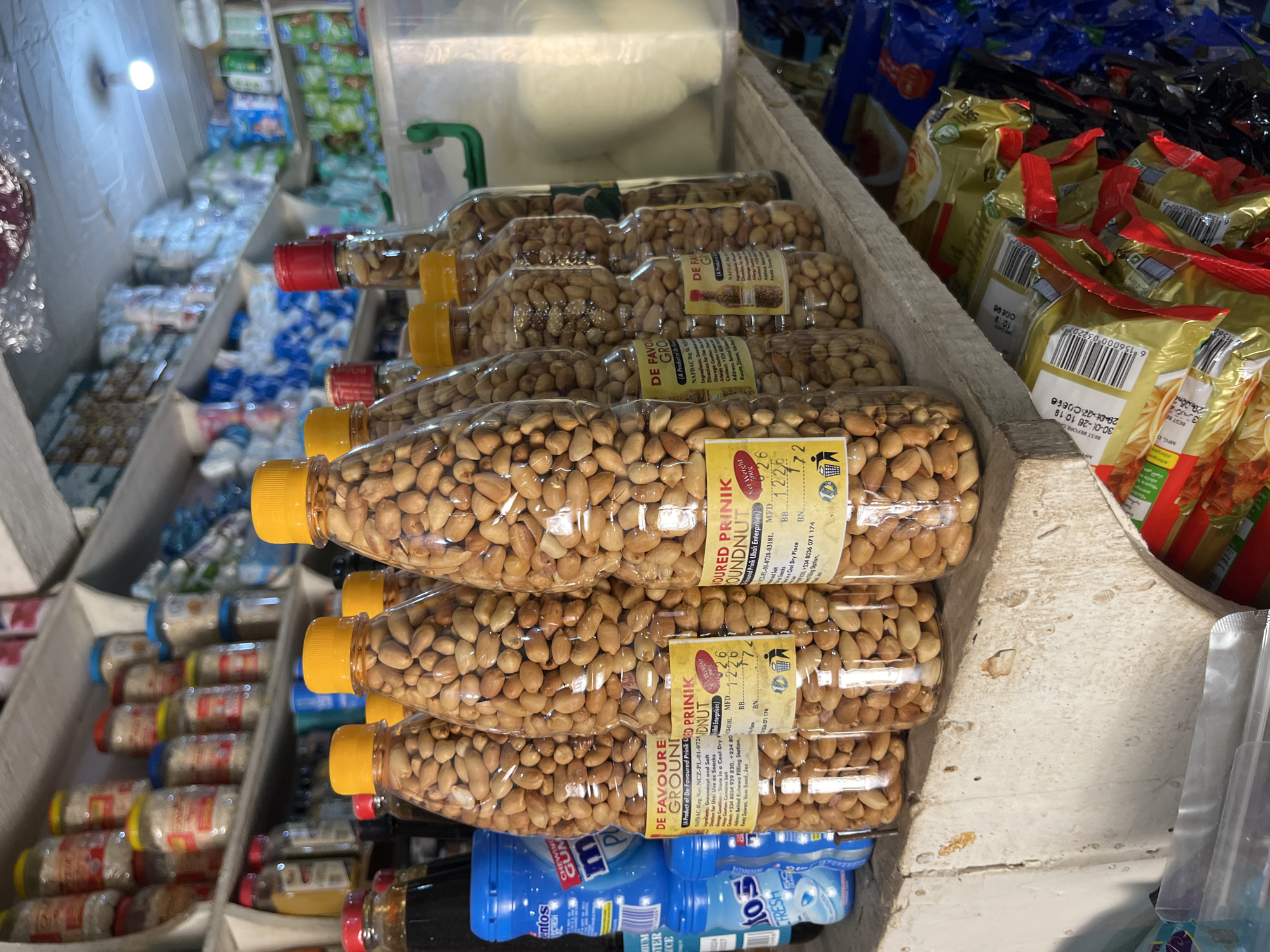 This image depicts a retail display in a shop in Bukuru, Nigeria, featuring shelves stocked with various food items. In the foreground, several clear plastic bottles filled with peanuts are prominently displayed on a wooden shelf. These bottles have yellow and red caps and some have yellow labels with text that includes "DE FAVOURED GROUNDNUT" and production details. To the left of the peanut bottles are blue plastic containers, one labeled "Mentos CHEWING GUM". Further back, shelves are lined with jars of what appear to be food products, some with red, blue, and yellow lids. The background reveals more shelving with packaged goods, including what looks like sachets of food or snacks. The overall environment suggests a small, possibly local, grocery or convenience store. The lighting is bright, illuminating the products on display.