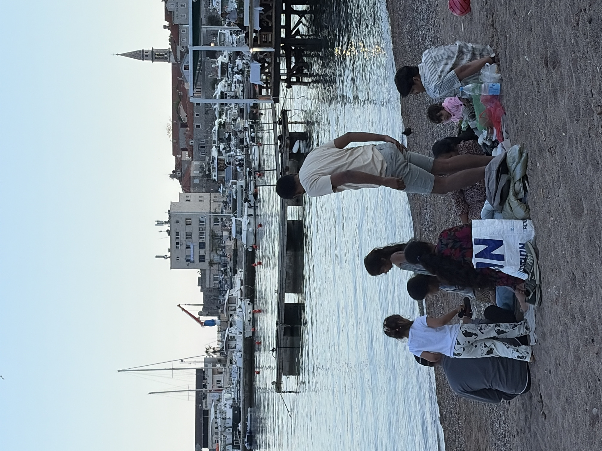 The image depicts a group of people on a beach in Budva, Montenegro. A family appears to be on the coast next to a harbor filled with yachts and other boats. There is a pier or dock-like structure extending into the water. Buildings are visible in the background, including a distinctive church tower that is a landmark of Budva's Old Town. The people are facing the water and appear to be engaged in an activity on the beach.