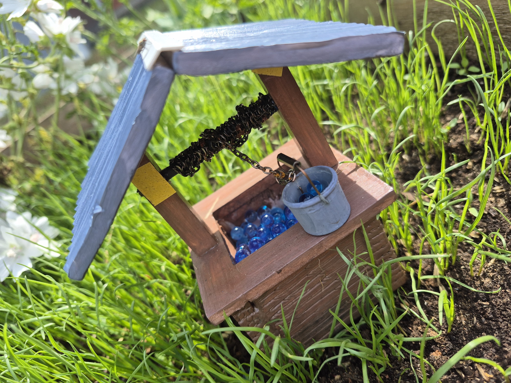 This is a close-up, eye-level shot of a miniature wishing well set amongst lush green grass and soil. The wishing well is constructed from wood, with a brown, textured base resembling logs. It features a slanted, blue roof supported by two wooden beams, each adorned with a small yellow square. A chain mechanism, appearing to be made of dark metal, is suspended from the roof, with a tiny, silver bucket hanging from it. The bucket is filled with bright blue glass beads, which also fill the well itself. In the background, out-of-focus white flowers and more green foliage are visible, suggesting an outdoor setting. The lighting indicates it is daytime, likely sunny, given the bright highlights and shadows. There is no visible text. The scene evokes a sense of whimsy and miniaturization.