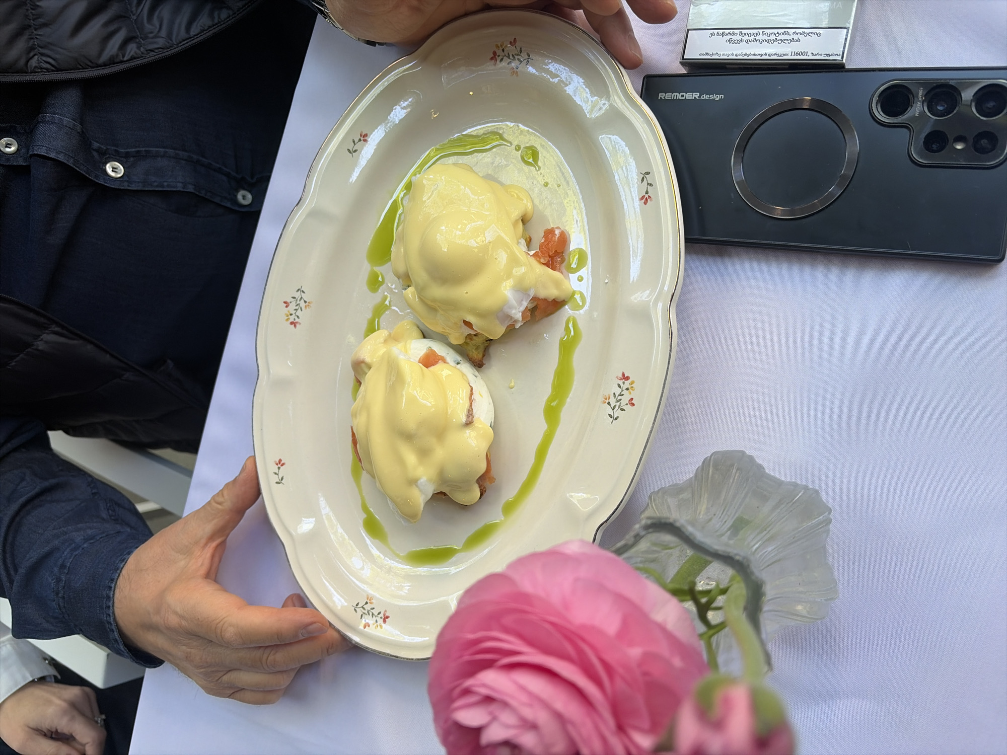 Here is a description of the image:
The image shows a table setting in what appears to be a cafe or restaurant setting. The food dish features two eggs benedict served on an ornately decorated plate with a gold trim. The eggs are topped with a yellow sauce, likely hollandaise, and garnished with a streak of green sauce. The plate is being held by two hands. A pink flower in a vase is positioned near the plate.
To the right of the plate is a black smartphone, partially obscured by a pack of cigarettes. The phone is labeled "REMOER.design," and the cigarette pack has text written in Georgian. The text indicates the location of the city of Tbilisi, Georgia. The table is covered with a white tablecloth.
In the background, a person is wearing a dark-colored jacket.