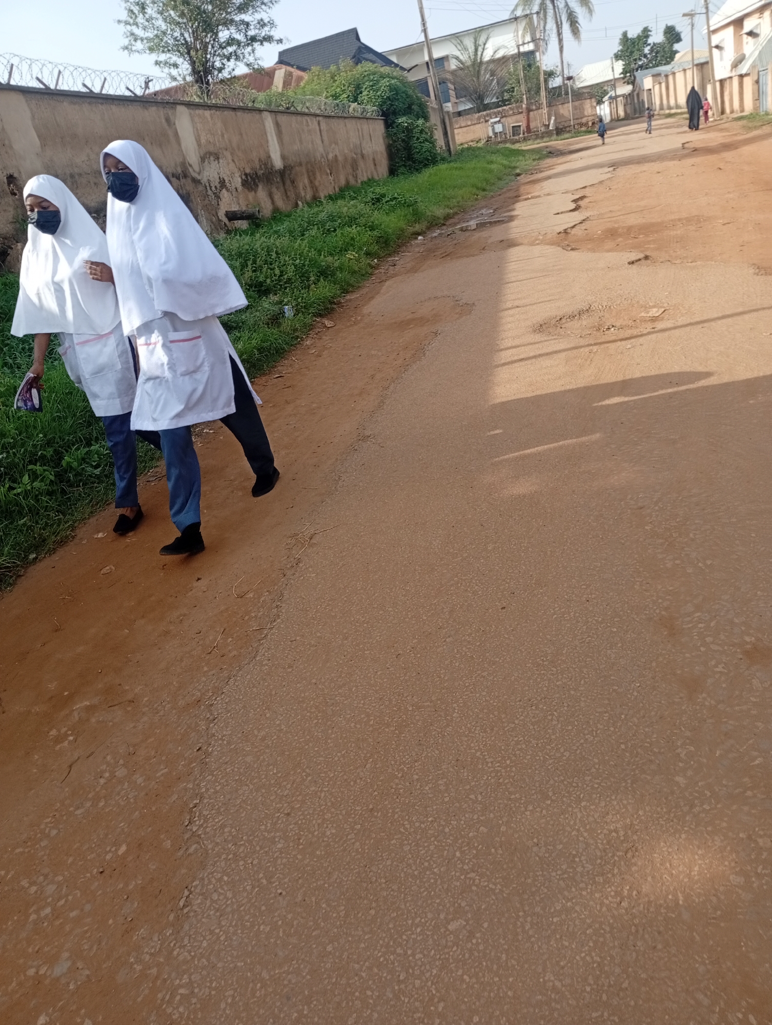 The image depicts a street scene in Rafin Par, Nigeria. Two individuals, appearing to be young women, are walking on a dirt and asphalt road. They are both wearing white hijabs and what seem to be white lab coats over dark blue trousers. They are also wearing face masks. The individual on the left is holding a purple and white object. The road is bordered by a low wall and overgrown grass on the left. In the background, there are buildings and trees. The road itself is in a state of disrepair with visible cracks and potholes. Shadows from unseen objects stretch across the road, indicating a bright sunny day. Other people and buildings are visible further down the street in the distance, suggesting a populated area.