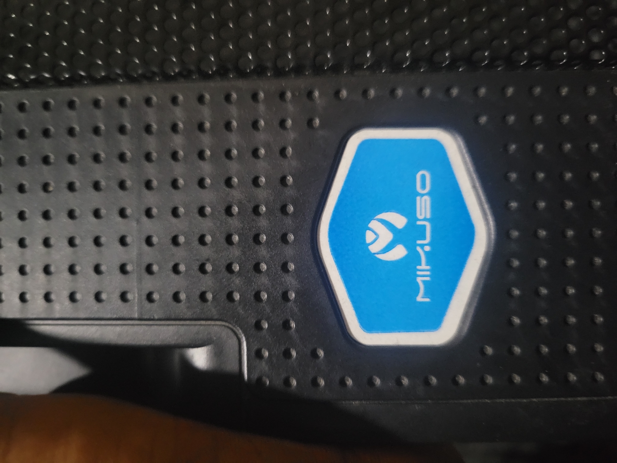 A close-up shot shows a black textured surface with a blue and white hexagonal emblem in the center. The emblem has a white border and contains a white logo with a stylized "M" and the word "MIKUSO" written below it in white capital letters. The textured surface is composed of small, circular indentations arranged in a grid pattern. The overall lighting suggests an indoor setting, possibly a product shot or a detail of an electronic device. There are no discernible elements that would place this image within a specific city or location.