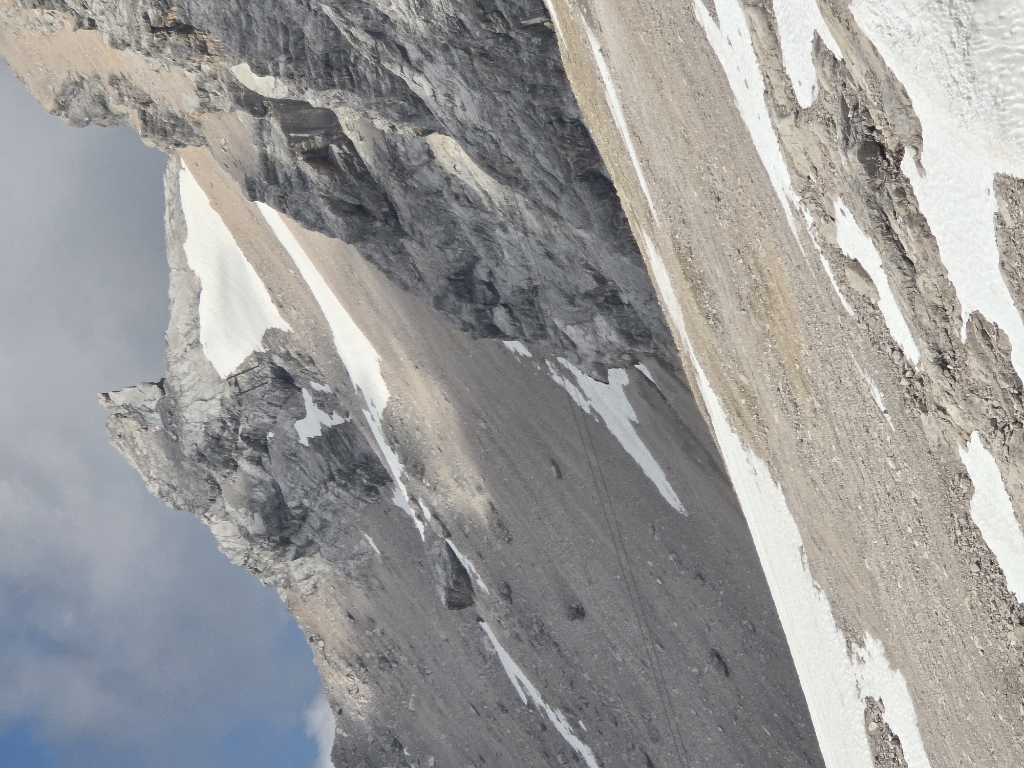 The image depicts a steep, rocky mountainside with patches of snow, characteristic of an alpine environment. The scene is dominated by shades of gray and brown rock, interspersed with stark white snow. The texture of the rock varies, showing layers and fissures, suggesting a rugged and weathered formation. Some areas are covered in scree, with smaller stones and pebbles.
The perspective is from above, looking down onto the slope. The angle of the image suggests a high vantage point, possibly from a helicopter or a higher point on the mountain itself. The lighting indicates it is daytime, with shadows cast by the terrain, but the overall light is somewhat diffused, possibly due to overcast conditions or haze. The presence of clouds in the lower left corner further supports this.
There are subtle details suggesting human activity. Faint, dark lines, possibly trails or ropes, can be observed on some of the scree slopes. In one of the cropped images, a tiny black speck on the slope might be a person, but it is too indistinct to confirm. The scale of the landscape dwarfs any potential human presence, emphasizing the grandeur of the mountains.
The context of Garmisch-Partenkirchen, Germany, places this scene in the Bavarian Alps, a region known for its dramatic peaks, hiking trails, and winter sports. The combination of rock and snow is typical for mountainous areas in this region, especially during transitional seasons like spring or autumn, or at high altitudes where snow persists year-round.
There is no visible text in the image. The overall mood is one of natural grandeur and a sense of remoteness, highlighting the raw beauty and imposing scale of the mountain landscape.