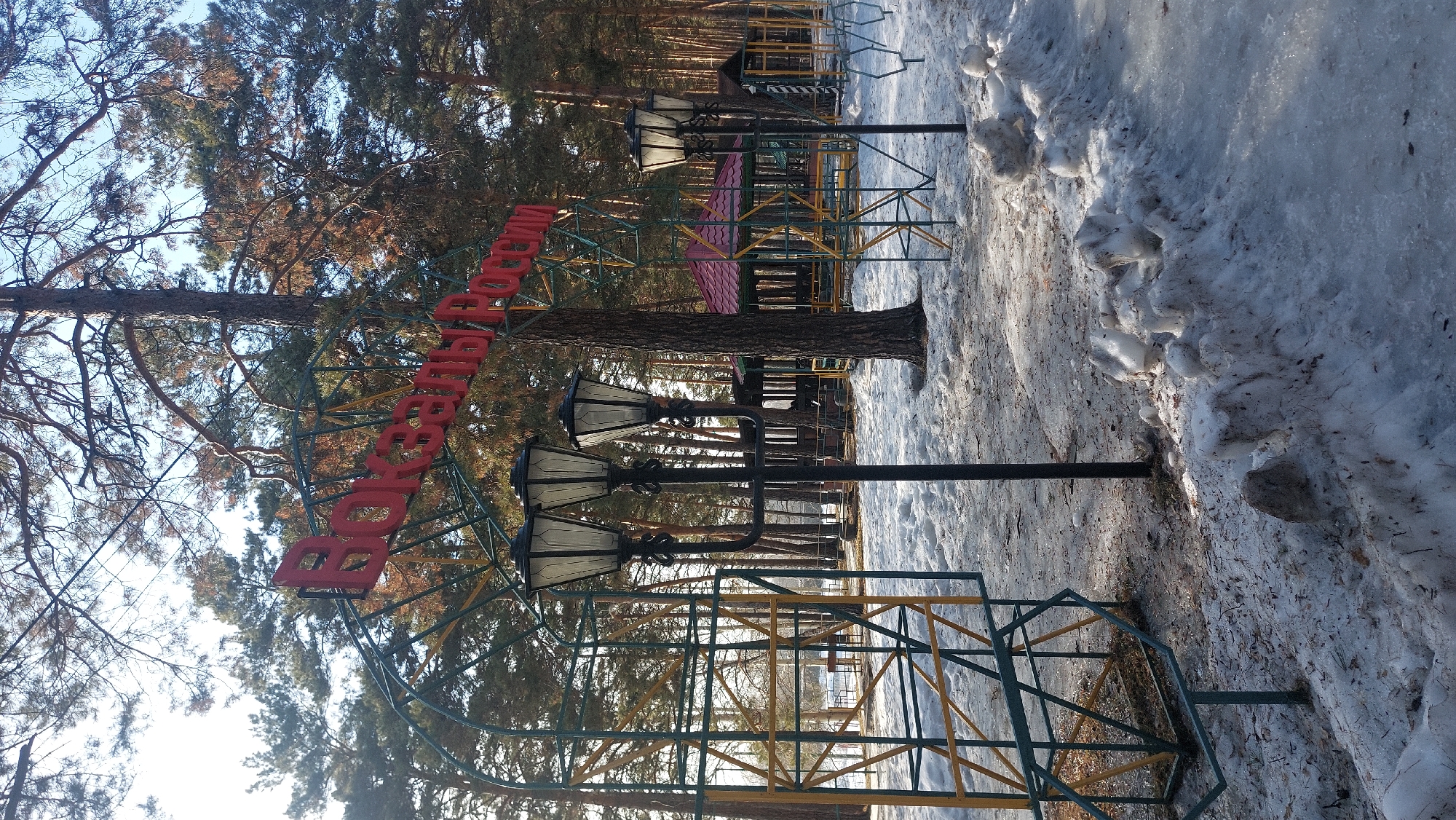The image depicts the entrance to a park in Novosibirsk, Russia. The sign "Березовая роща" (Berezovaya Roscha - Birch Grove) is displayed in red capital letters on a metal arch structure. Several vintage-style street lamps are positioned around the sign. The surrounding environment includes bare trees, snow-covered ground, and a playground structure visible in the background.