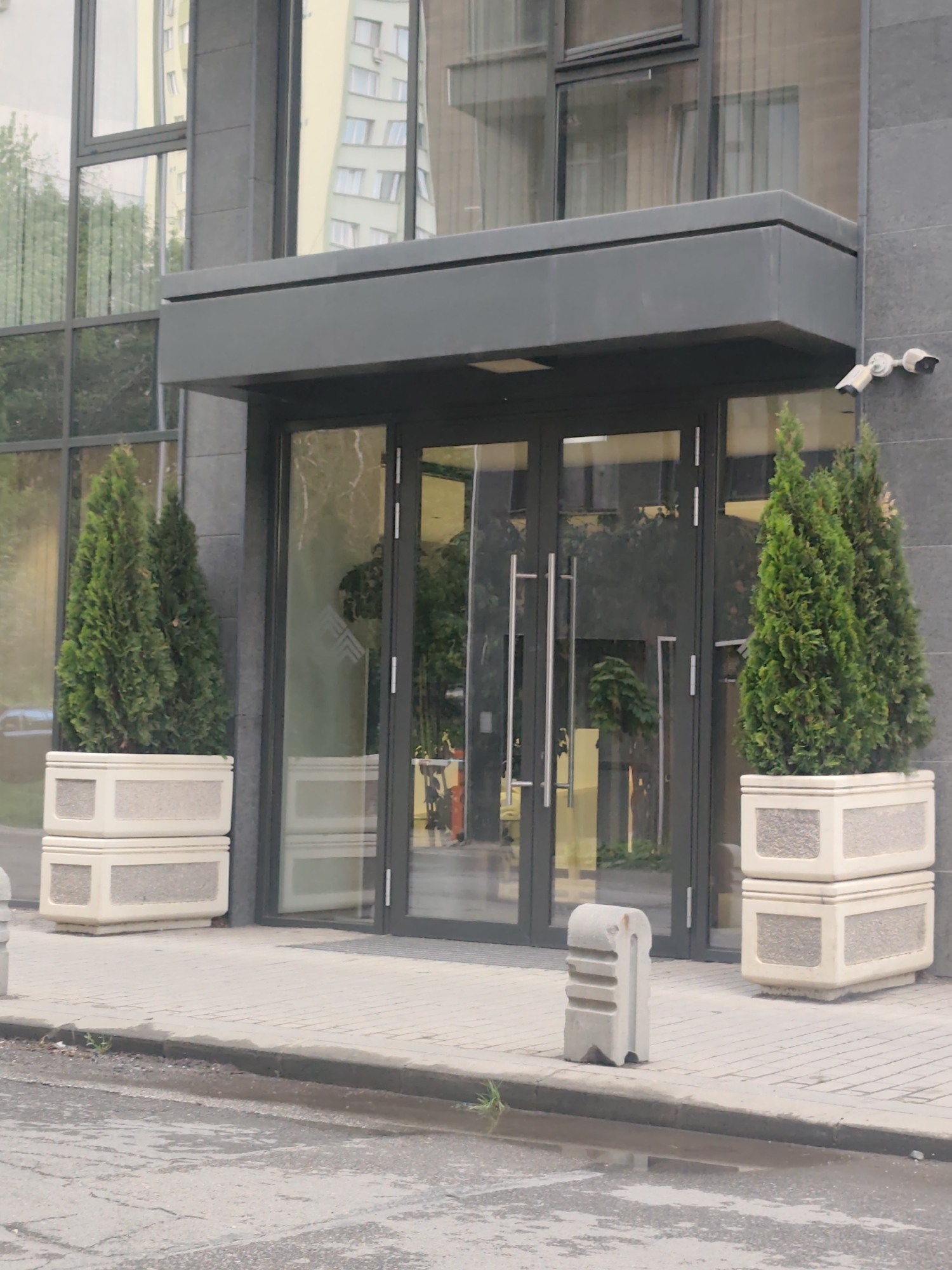 The image captures the entrance of a modern building in Sofia, Bulgaria, characterized by its sleek, functional design. The central focus is a set of dark grey-framed glass double doors, providing a transparent view into the building's interior, which reflects indistinct shapes and possibly artificial light. Above the entrance, a substantial dark grey awning or canopy extends outwards, providing shelter.
Flanking the entrance, two tall, conical coniferous plants, likely Thuja or Juniper, are neatly arranged in stacked, light-colored concrete planters with a coarse aggregate finish. These plants add a touch of natural greenery to the otherwise stark architecture.
The building itself features large, dark-framed glass windows that reflect the surrounding environment. Prominently visible in the reflections are parts of a light-colored, multi-story building with numerous windows, suggesting a dense urban setting. Green trees are also reflected in the glass, indicating nearby vegetation. The building's facade is clad in dark grey, possibly stone or composite panels, contributing to its contemporary aesthetic. Two white security cameras are mounted on the wall to the right of the entrance, monitoring the area.
In front of the entrance, a neatly paved sidewalk made of light grey pavers leads to a street or road. A small, light grey concrete bollard with carved horizontal lines stands on the edge of the sidewalk, acting as a barrier. The street appears damp with several puddles, suggesting recent rainfall or wet conditions. The time of day appears to be daytime, with soft, diffused lighting.
No discernible text is visible in the image. A faint, stylized double-arrow symbol pointing upwards is visible on one of the glass doors. The overall impression is one of a clean, well-maintained commercial or residential building in an urban environment.