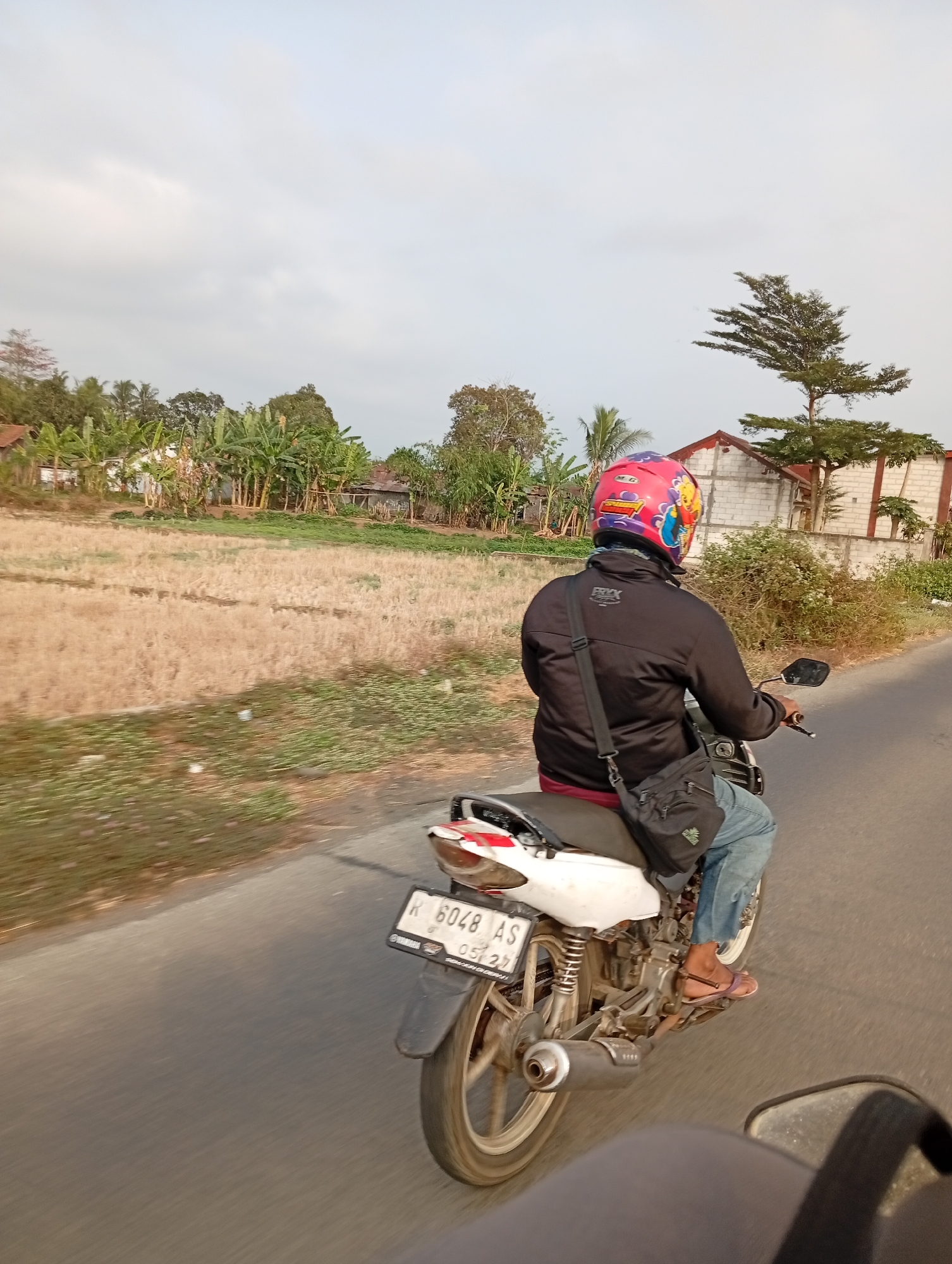 A person rides a motorcycle on a paved road in a rural area. The person is wearing a black jacket, blue jeans, and a pink helmet decorated with cartoon characters. A black bag is slung across their shoulder. The motorcycle is white and has a license plate with the letters "R 6048 AS". The environment is characterized by dry, yellow fields on the left side of the road and vegetation, trees, and houses on the right. The sky is overcast with light gray clouds. The image appears to have been taken from another moving vehicle.
