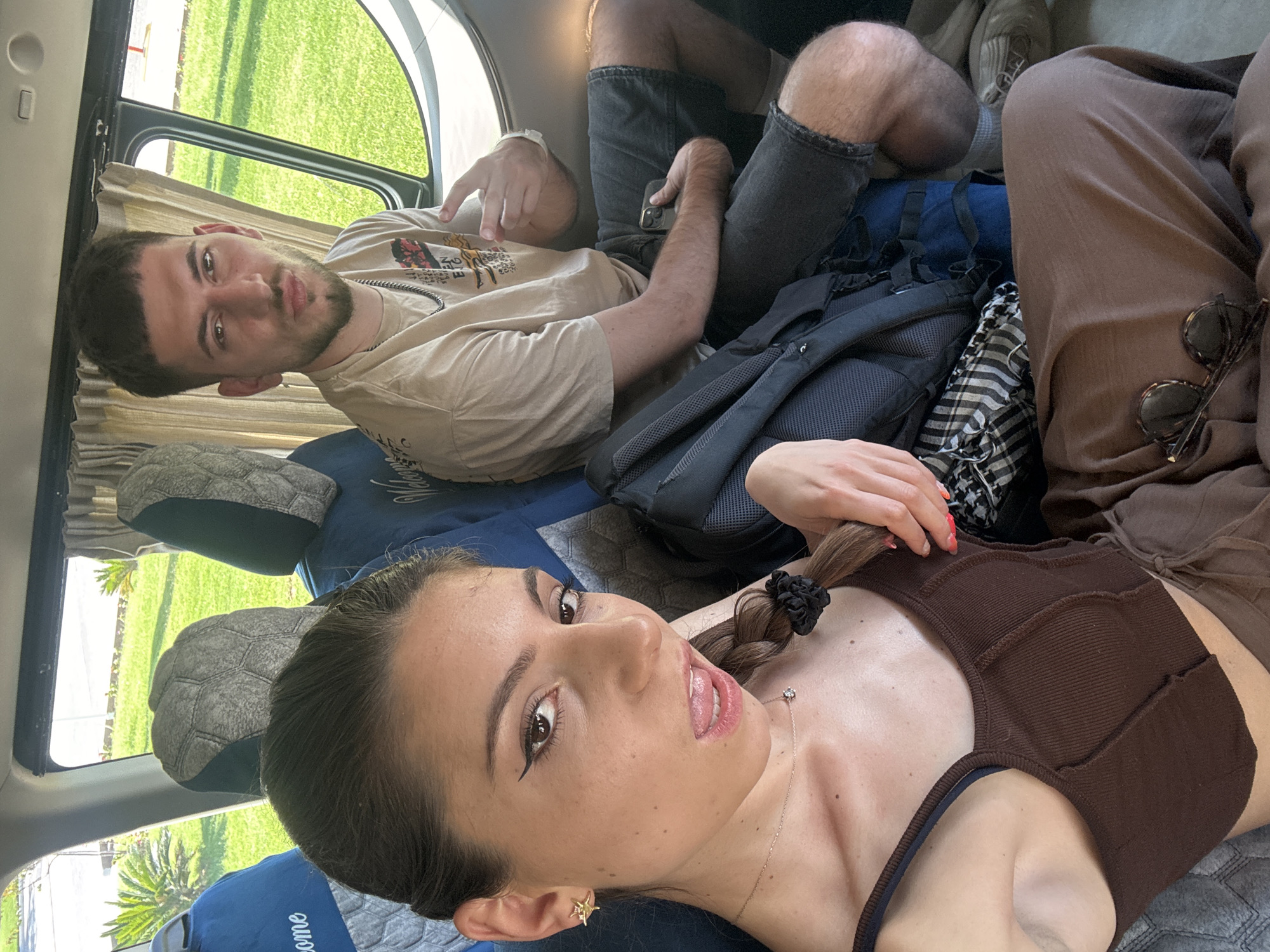 Here is a detailed description of the image:
Two young adults are lying in the back of a vehicle, possibly a campervan, in Unknown, Unknown. The woman is in the foreground, lying on her back, wearing a brown top and brown pants. Her hair is dark and pulled back in a ponytail with a black scrunchie. She has eyeliner on and wears a necklace. Her mouth is slightly open. Sunglasses are positioned next to her on her pants.
Behind her, a man is also lying down, wearing a tan t-shirt and gray shorts. He has short dark hair and a beard. He is pointing with his right hand. A black backpack is situated between them.
The interior of the vehicle is visible, with blue cushions and window with curtains. Outside the window, green vegetation is visible.