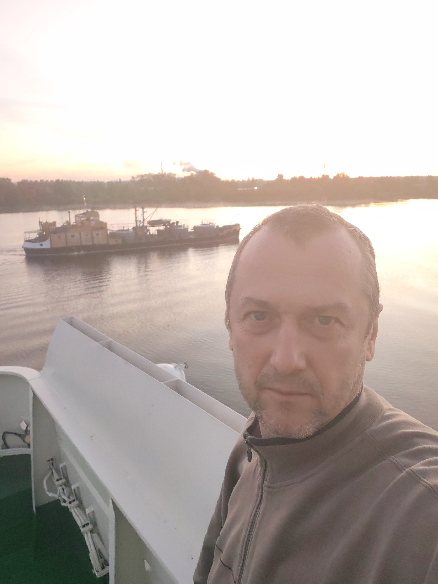 A man takes a selfie on a ship. Behind him is a river with another boat on it. The far shore has buildings and trees. The time of day appears to be sunrise or sunset, with the sky reflecting on the water. The location is Unknown, Unknown.