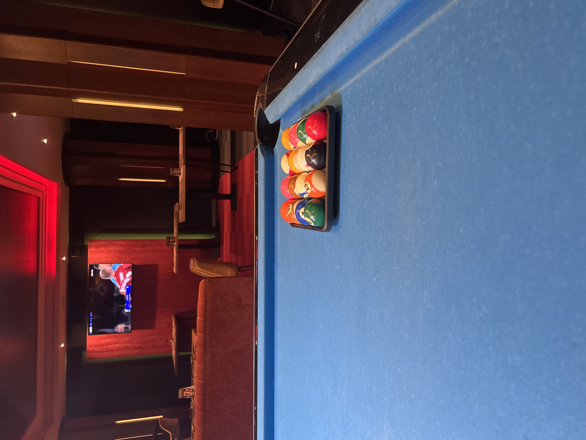 The image depicts a pool table with a set of billiard balls racked at one end. The table is covered in blue felt and has a dark-colored rail. A black triangular rack is holding the balls. The background shows a dimly lit bar or lounge area with red lighting, a television screen displaying a sports game, and various seating options including booths and chairs. The location appears to be a bar in an unspecified city. Without additional data, the city of Sofia, Bulgaria, can not be determined.