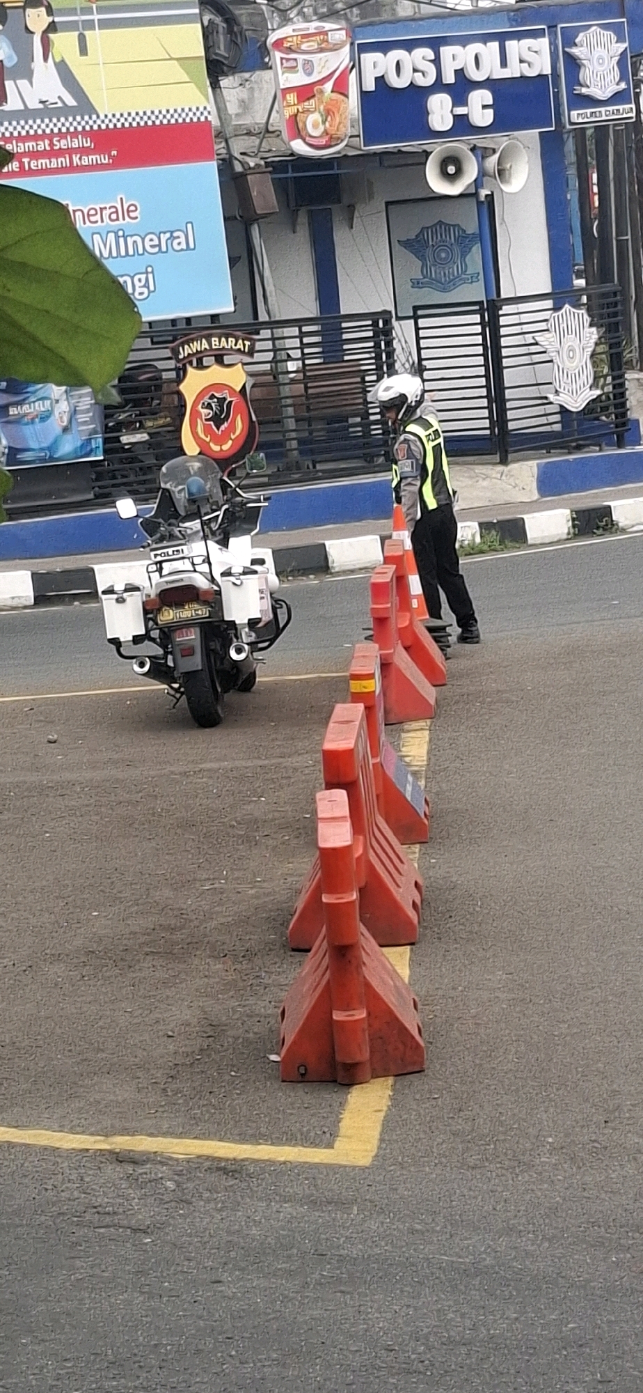 At a police post in Cianjur, Indonesia, a police officer wearing a white helmet and a yellow vest stands near traffic barriers. A white police motorcycle is parked behind the barriers. The police post has a sign reading "POS POLISI 8-C". A large green leaf obscures part of the scene.
