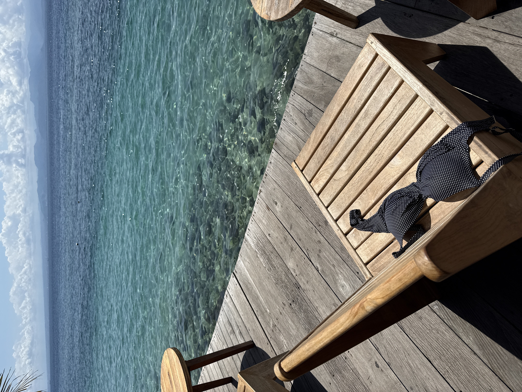 View from a wooden deck overlooking turquoise water, presumably in Nusa Penida, Indonesia. A black and white polka-dot bikini top rests on a wooden deck chair. The deck extends over clear water, revealing the sea floor. The horizon shows a distant landmass.