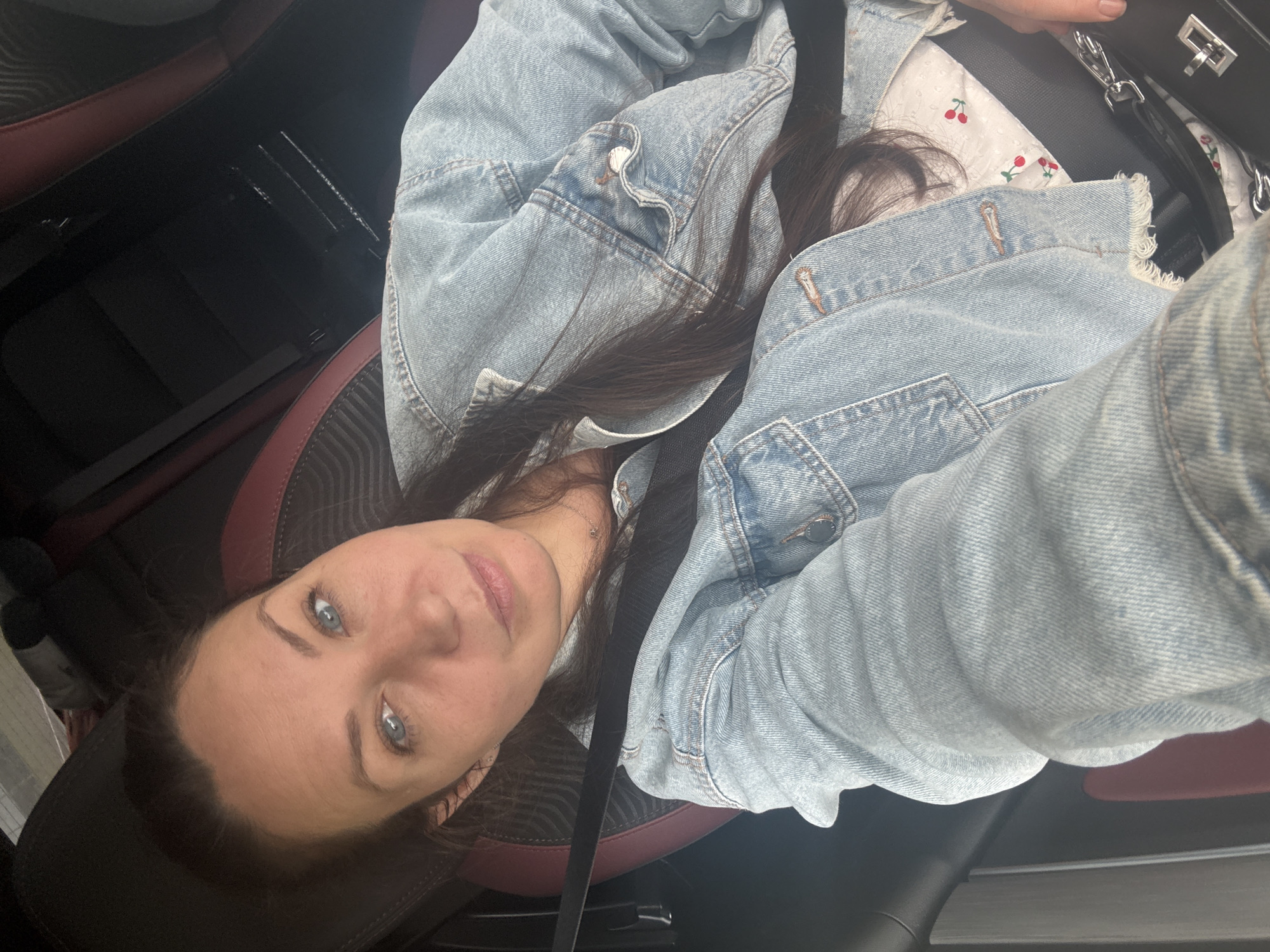 This media file is a selfie taken by a woman from inside a car. She is wearing a light blue denim jacket over a white shirt with a cherry pattern. Her long, dark hair is visible. The woman has striking blue eyes and fair skin with a light dusting of freckles. She is looking directly at the camera with a neutral expression.

The car's interior is visible, with dark seats that appear to be leather with red accents. A black seatbelt is across her body. A black handbag with silver hardware is also visible. The lighting suggests it is daytime, possibly overcast or in a shaded area, as there are no harsh shadows.

The image is taken from a slightly high angle, looking down at the woman. The focus is on the woman, with the car interior serving as the backdrop. There is no visible text in the image. The overall impression is a casual, everyday moment captured while in a vehicle.