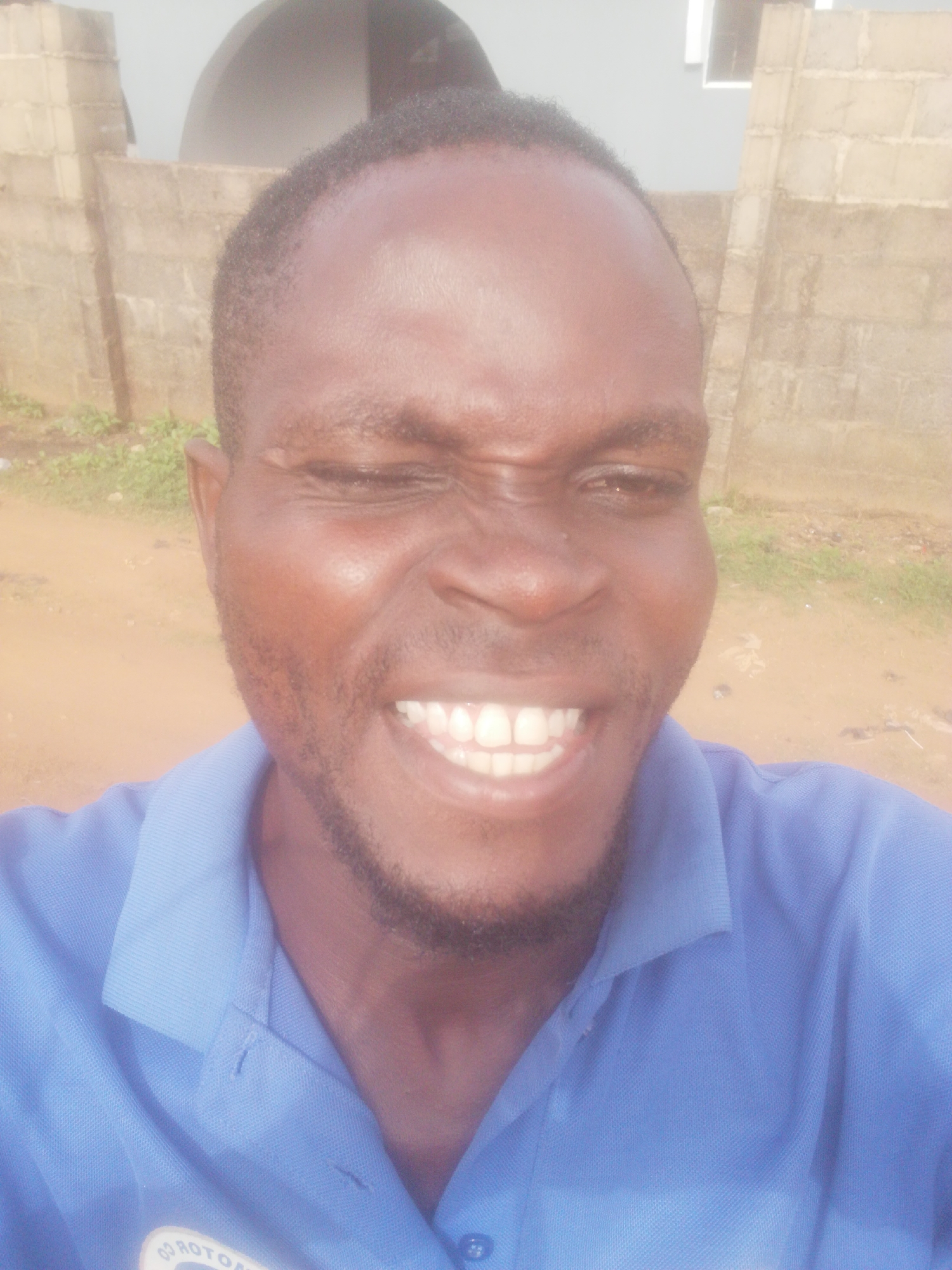 A close-up shot shows a man with dark skin, short black hair, and a beard, smiling widely with his eyes squinted shut. He is wearing a blue polo shirt. Behind him, there is a concrete block wall and a patch of green vegetation. The location appears to be outdoors in an urban or suburban area.