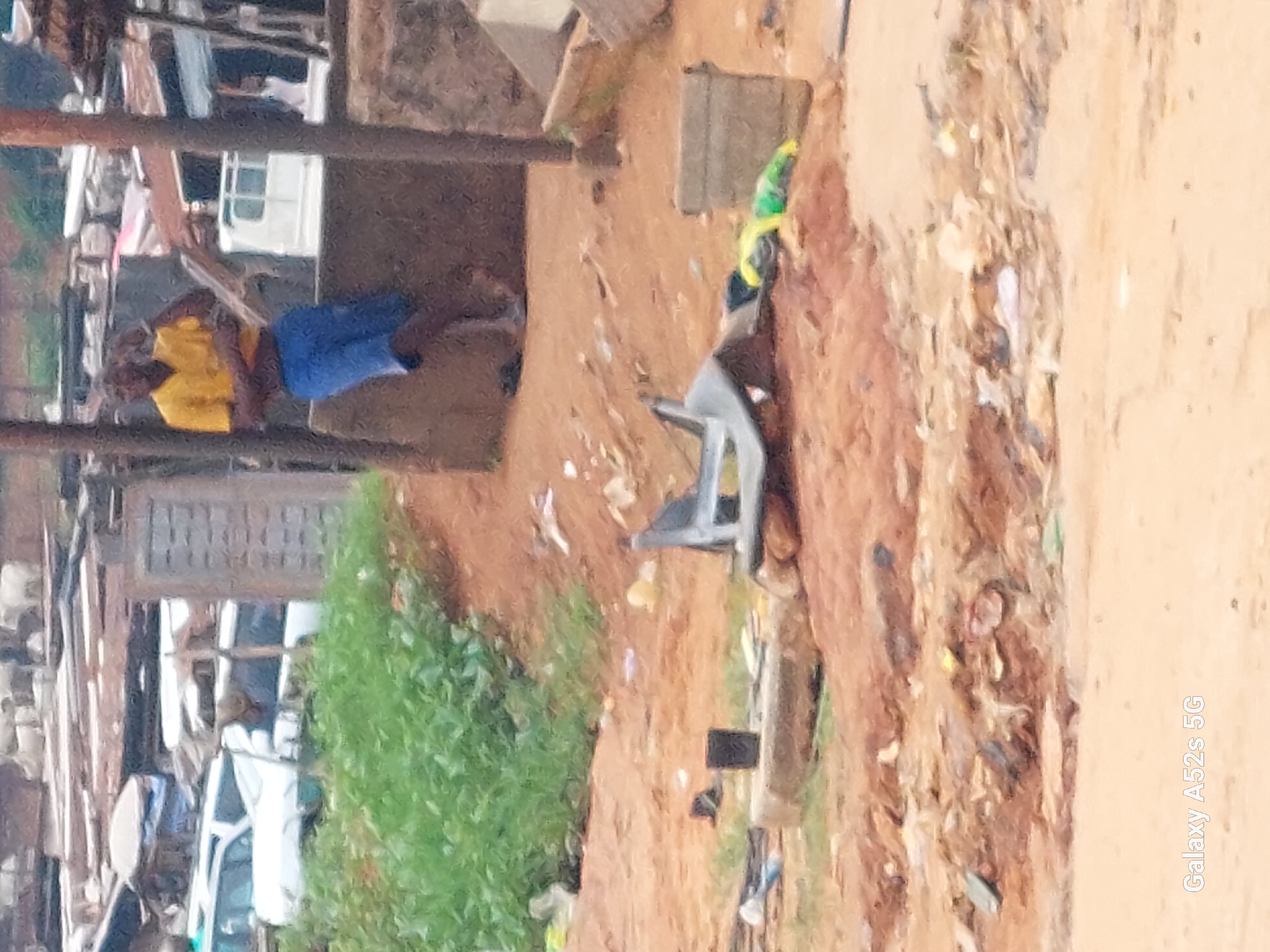 In Awkuzu, Nigeria, a young man in a yellow t-shirt and blue shorts stands leaning against a brown, textured wall. The environment appears to be an informal settlement or a semi-developed area, characterized by rough terrain and scattered debris. Behind him, a white van is visible, along with other structures suggesting an open-air market or residential area with makeshift constructions. The ground is predominantly red earth, with patches of green vegetation in the background. Scattered on the ground are various objects, including what appears to be a discarded plastic chair, and some plastic bags. The overall impression is one of a utilitarian and somewhat untidy urban setting.