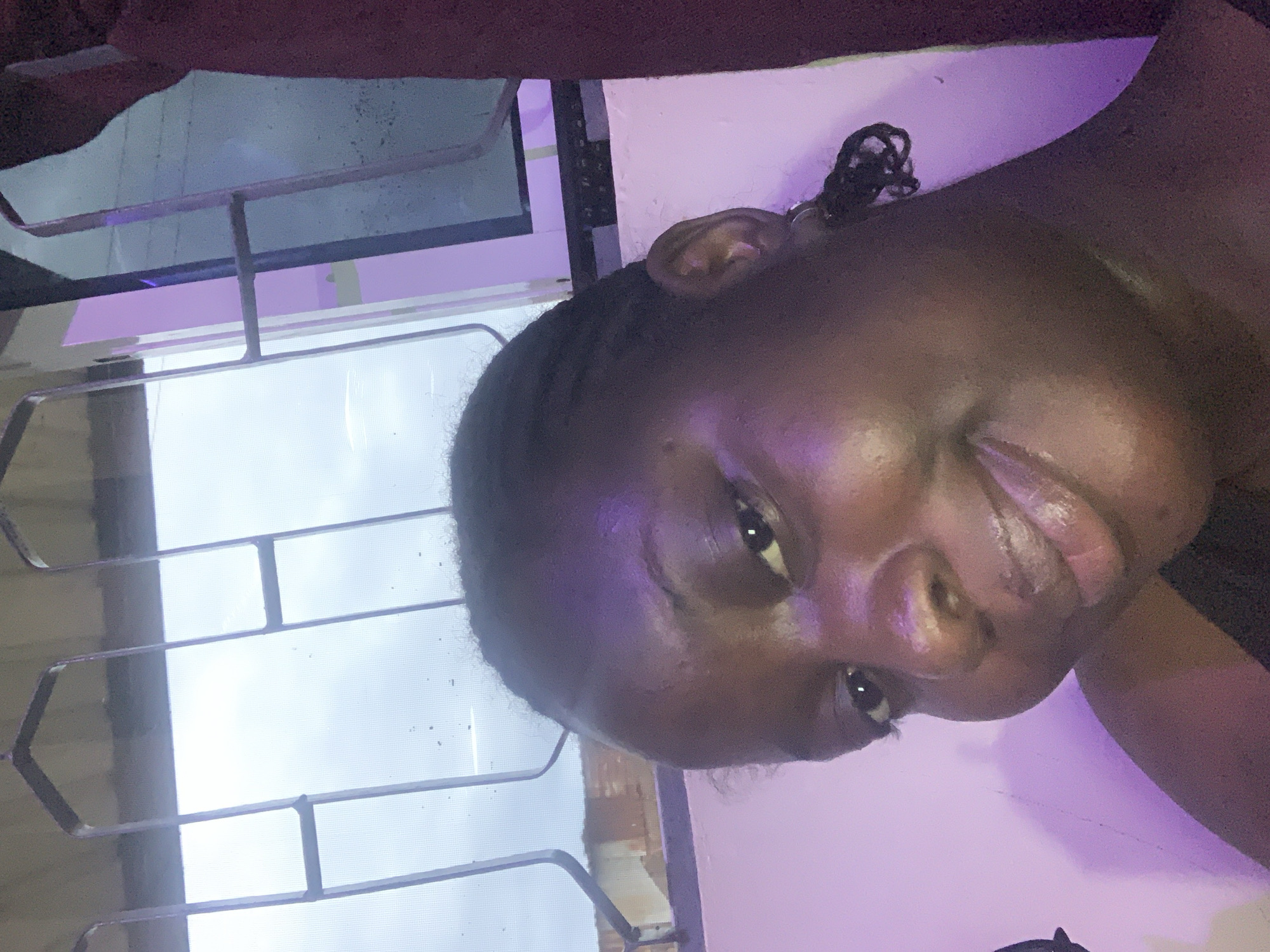 A selfie of a young Black woman taken from a low angle. She has braids and is smiling directly at the camera. Her skin is dark and appears dewy, with purple lighting reflecting off her face. She is wearing a dark top. Behind her, a window with bars is visible, and beyond that, a cloudy sky. A burgundy curtain is on the right side of the frame. The background suggests an indoor setting with a window.