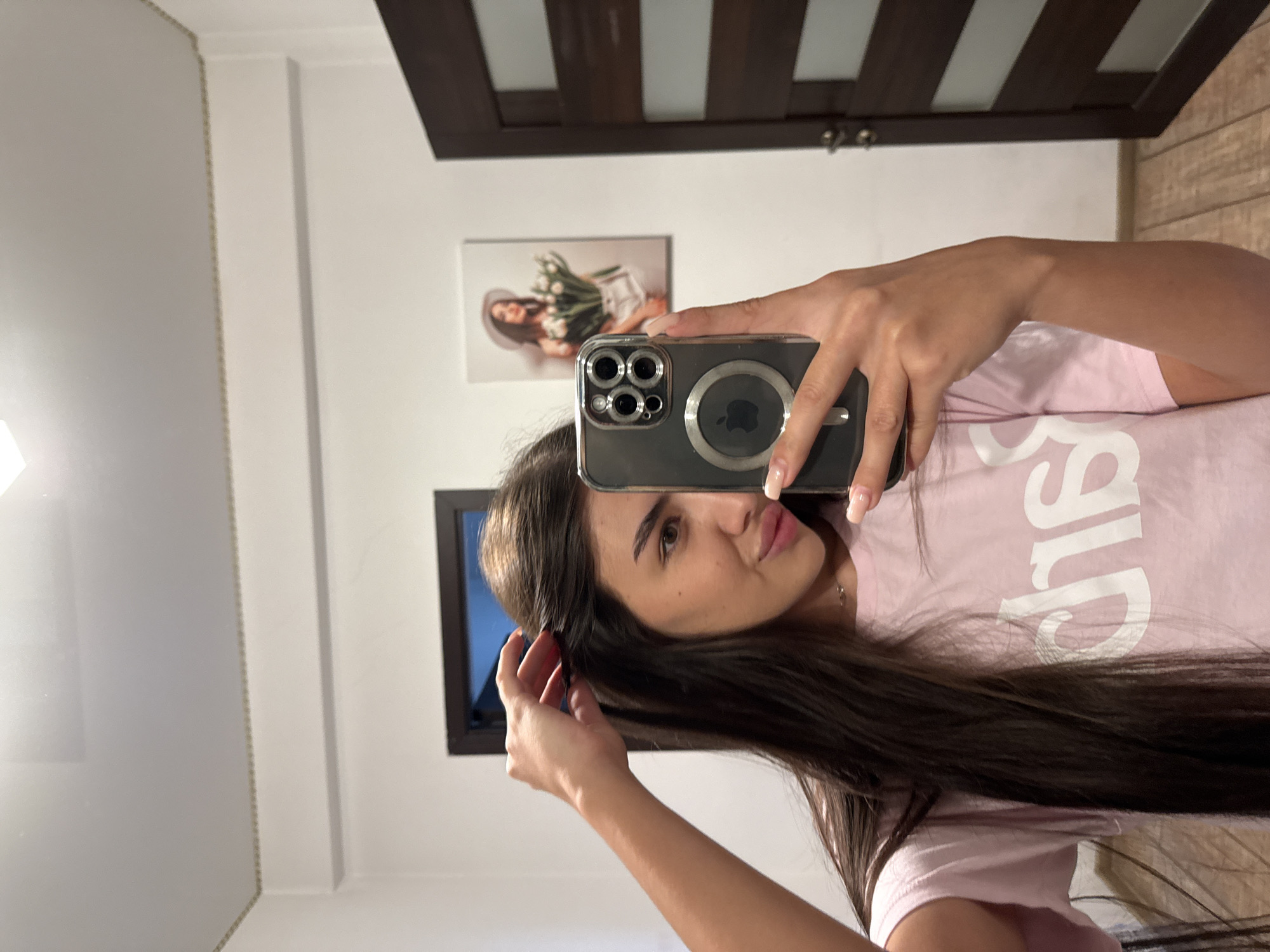 The image shows a young woman taking a mirror selfie in a room. She is wearing a pink t-shirt with white lettering. Her hair is long and dark brown, and she is touching it with her left hand. She is holding a smartphone in her right hand, which is also reflected in the mirror. In the background, there is a framed picture on the wall, a dark wooden door, and what appears to be wooden flooring. The room has white walls and a white ceiling with lighting fixtures. The overall setting suggests a domestic environment, likely a bedroom or hallway. The image does not contain any specific landmarks or contextual clues that would allow for the identification of its location within Chișinău, Moldova.