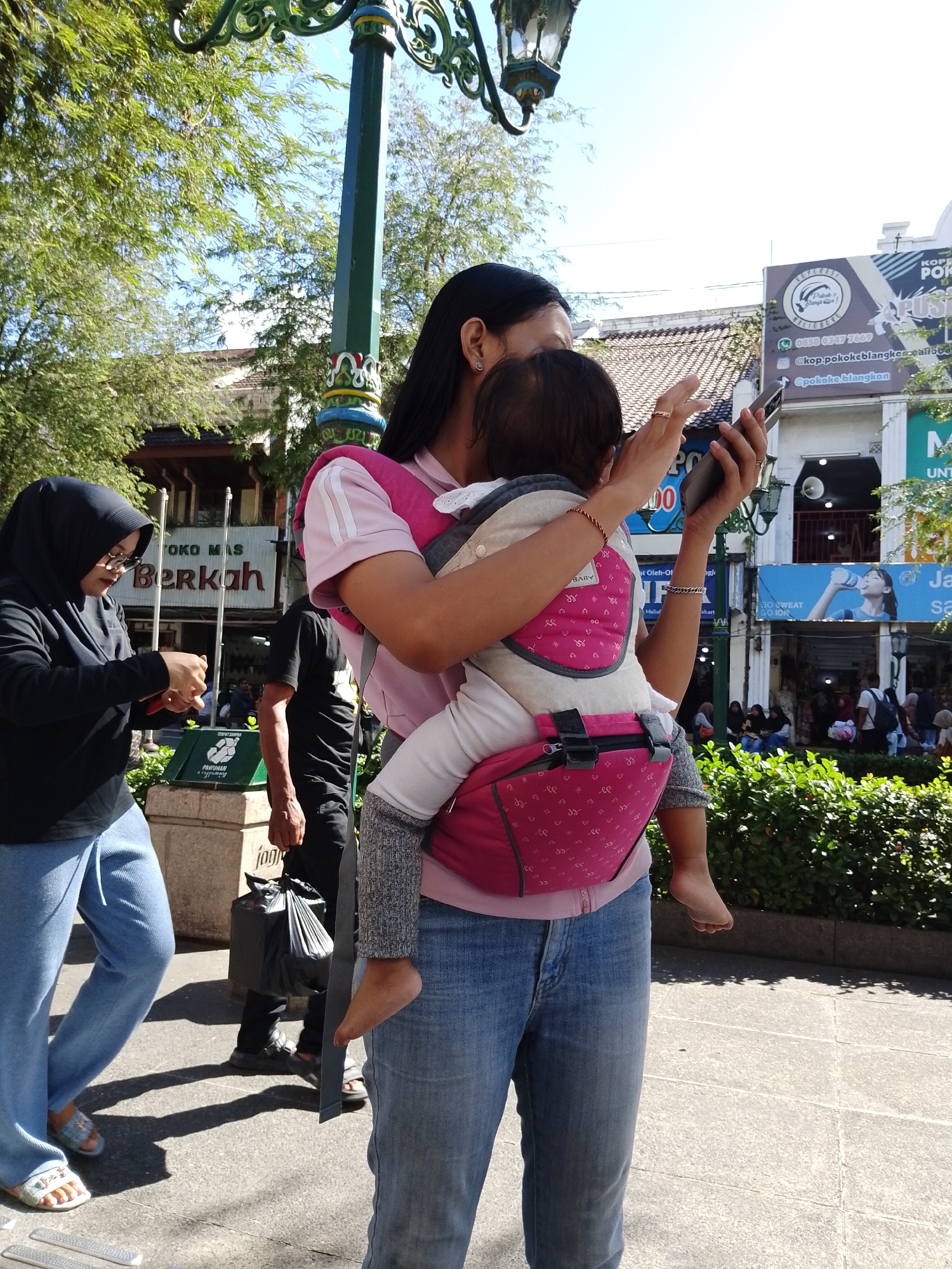 This image captures a bright, sunny day in Yogyakarta, Indonesia, likely in a busy urban area given the presence of shops and a street lamp. The primary subjects are people walking outdoors. In the foreground, a woman is carrying a baby in a pink and grey front carrier. She is looking at her phone, suggesting she might be navigating or communicating. The baby's small foot is visible, peeking out of the carrier.
To the left, another woman wearing a black hijab and glasses is also looking down, possibly at her phone or something in her hands. A man in a black t-shirt walks behind them, carrying a large black plastic bag. In the background, several other individuals are visible, some sitting down.
The scene is set along a paved walkway with some greenery and shops lining the street. Visible text includes "TOKO MAS Berkah" (Gold Shop Berkah), implying a commercial area. Other text on signs in the background relates to "KOPI POKOKE BLANGKON" (a coffee shop) and "GO SWEAT GO ION" (likely an advertisement for a drink or sports-related product). The overall atmosphere appears to be one of everyday life and commerce in a bustling Indonesian city.