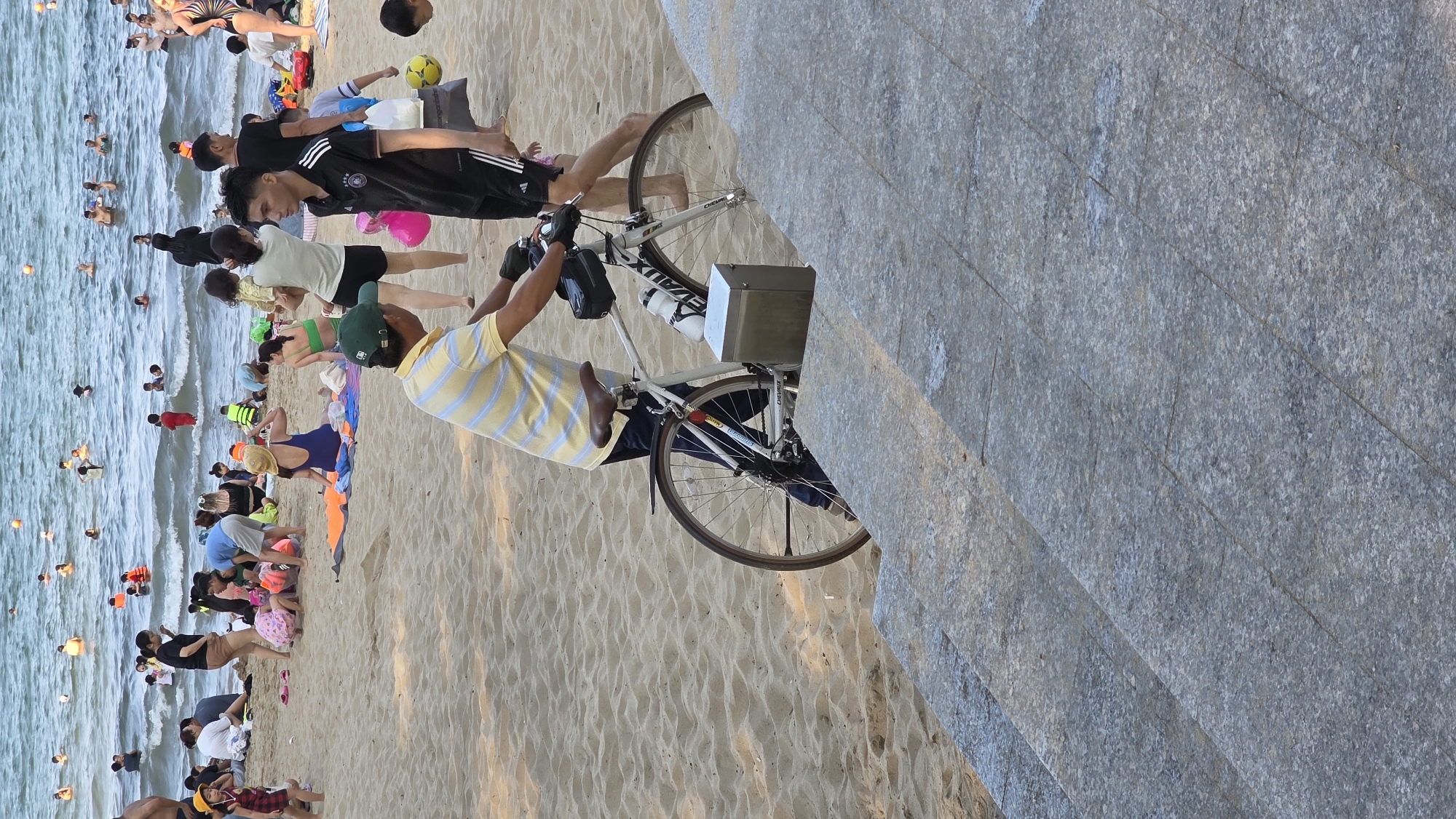 The image depicts a beach scene in Nha Trang, Vietnam. People are swimming in the ocean and relaxing on the sand. A man in a striped shirt stands next to a bicycle on the beach. Nearby, other people are standing and sitting on the sand. The background includes a concrete structure.