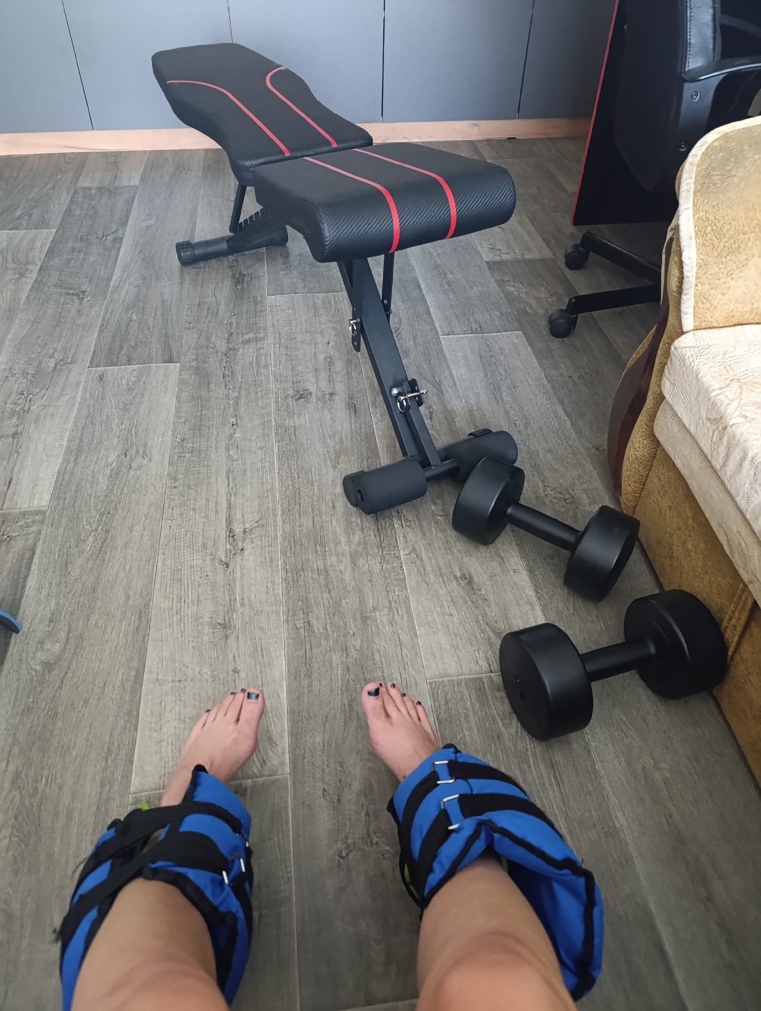 The image shows a home gym setup on a wooden floor. A black and red adjustable weight bench is positioned centrally, with two black dumbbells lying on the floor to its right. In the foreground, a person's legs are visible, wearing blue and black ankle weights. To the right of the bench, a portion of a tan sofa is visible, and behind it, a black office chair with wheels and part of a desk. The environment appears to be an indoor residential space. The specific location of Novokuybyshevsk, Russia, is not evident from the visual information provided in the image.