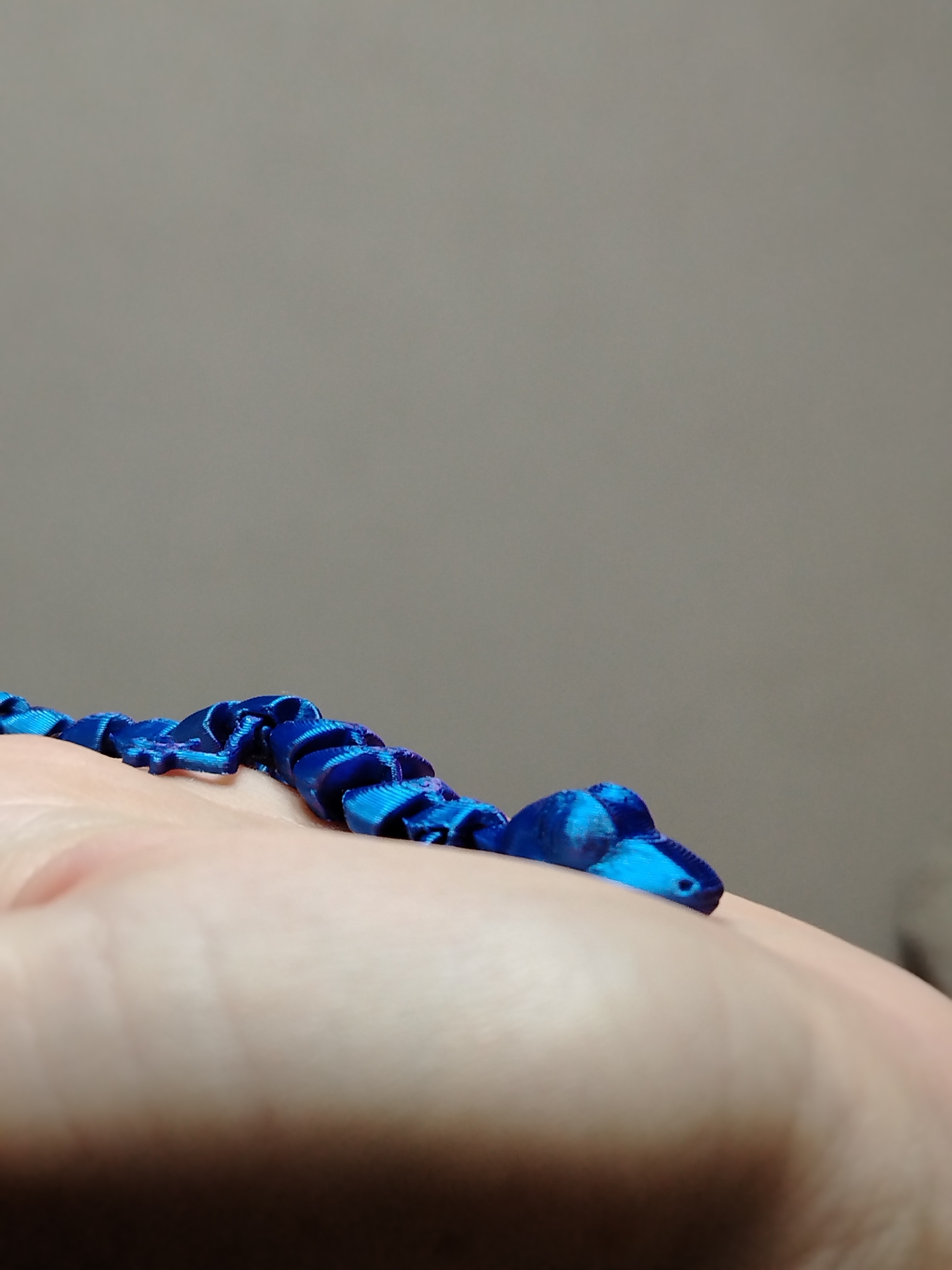 A close-up shows a flexible, articulated toy snake made of blue material, possibly 3D-printed or fabric. The snake rests on a human hand, with the background consisting of a plain grey or taupe surface. No other details, including the location in Izhevsk, Russia, are evident in the image.