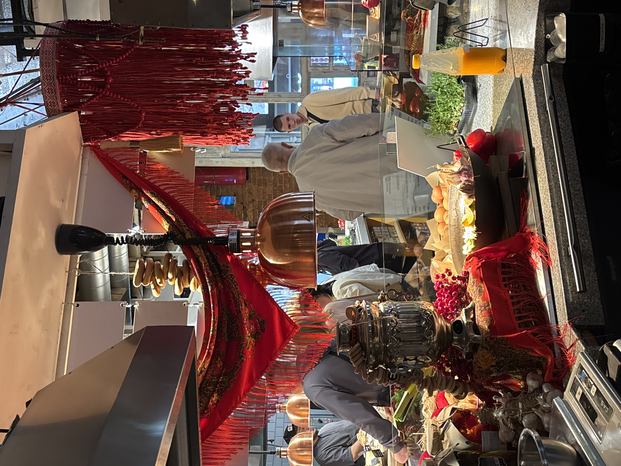 The image depicts a market stall in Saint Petersburg, Russia, featuring a traditional Russian samovar as a central decorative element. The stall is adorned with a vibrant red scarf with intricate patterns and fringes. Several people are visible, some interacting with the stall's offerings, suggesting a bustling atmosphere. Among the items on display are bagels, garlic, red berries, and a bottle of orange juice. The environment appears to be indoors, possibly a food hall or market, with brick walls and industrial lighting fixtures visible in the background.