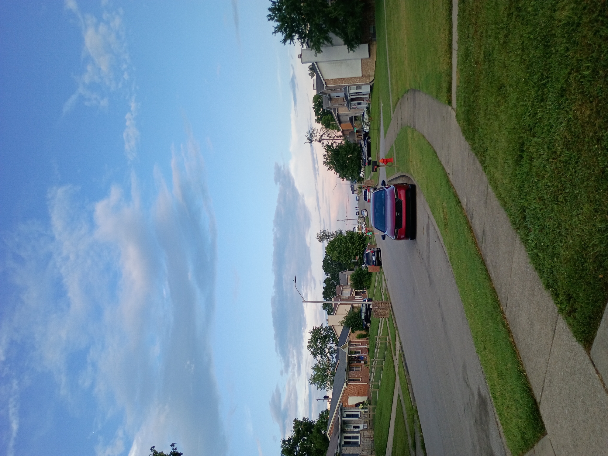 The image depicts a suburban street scene in Nicholasville, Kentucky, USA, under a partly cloudy blue sky. The perspective is from a sidewalk, looking down the residential street lined with single-family homes on both sides. Several cars are parked on the street and driveways, including a red sedan in the foreground, a black SUV further down the road, and other vehicles visible in the distance. The houses appear to be of various architectural styles, primarily brick and vinyl siding, with well-maintained lawns. A fire hydrant is visible near the curb in the middle ground. Trees are abundant along the street and in the yards, some with full foliage. The overall impression is a quiet, residential neighborhood in the late afternoon or early evening, indicated by the soft lighting and the pastel hues in the sky.