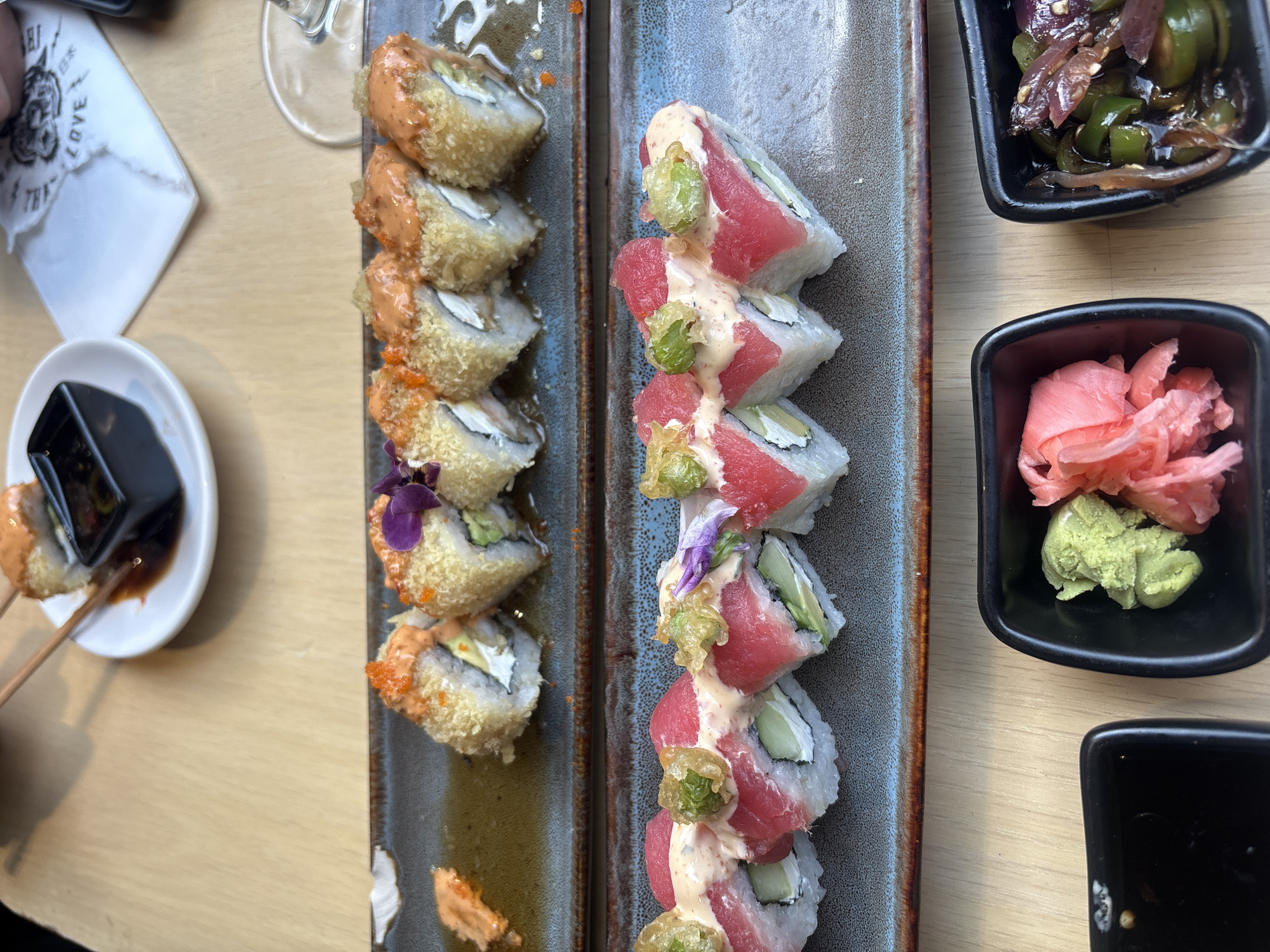 The image shows two plates of sushi on a wooden table. One plate contains fried sushi rolls topped with orange sauce and a small purple flower. The other plate holds sushi rolls with tuna, cucumber, and a white sauce, garnished with green dollops and purple flowers. Two small black bowls contain pickled ginger, wasabi, and a mix of green peppers and red onions. A small white plate with soy sauce and chopsticks is visible. The scene is possibly in a restaurant in Mexico City, Mexico, suggested by the presence of sushi, a popular international dish in the city's culinary scene.