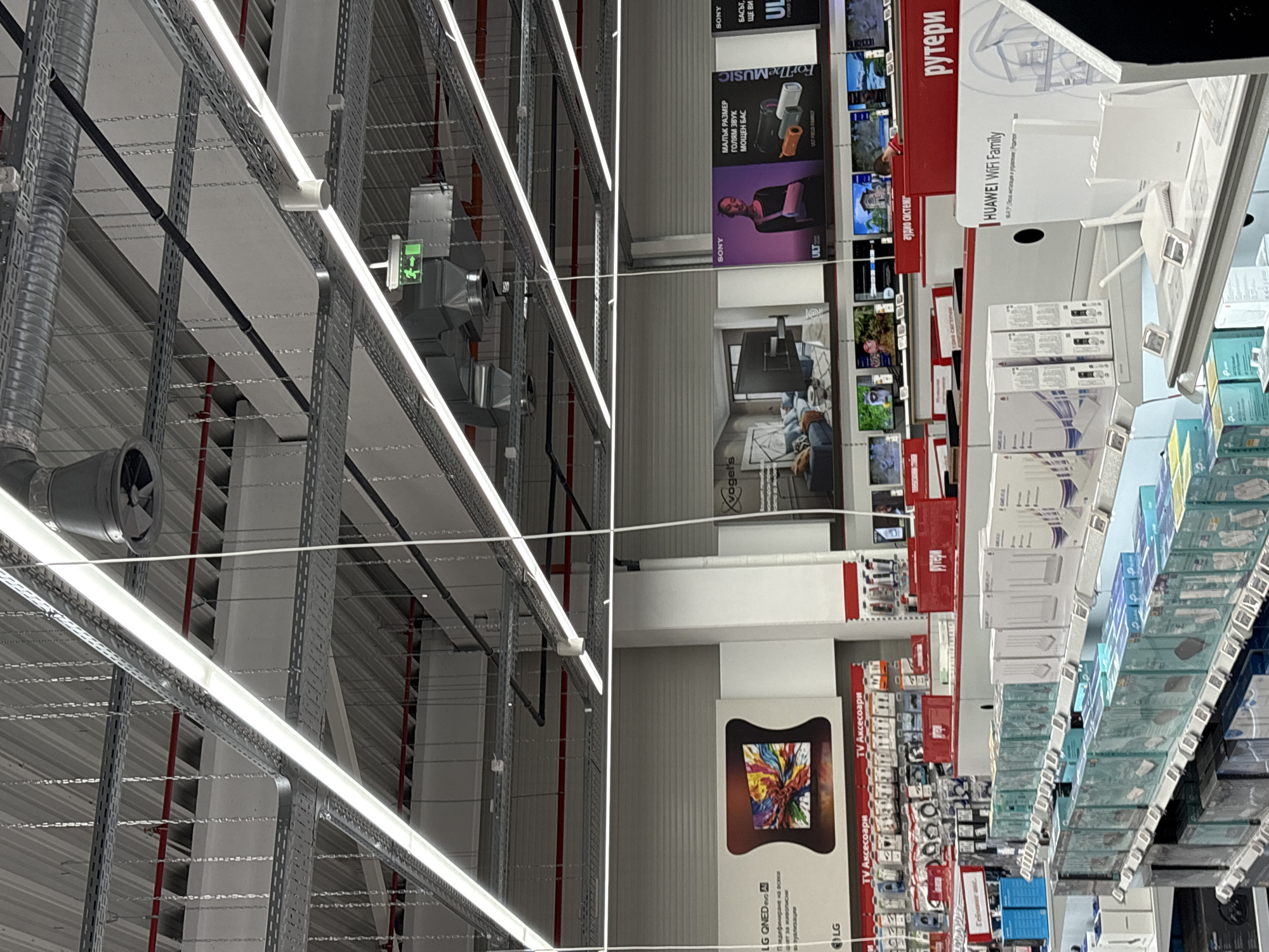 The image shows the interior of a retail store, likely in Lovech, Bulgaria based on the presence of Cyrillic text. The left side of the image features industrial ceiling elements including ductwork, lighting fixtures, and cable trays. The right side of the image displays retail shelves stacked with electronics products, including routers and accessories from brands like Huawei, TP-Link, and LG. Sony audio products and televisions are advertised. The overall environment suggests a store specializing in electronics, potentially a department store with an electronics section. There are no visible people in the frame.