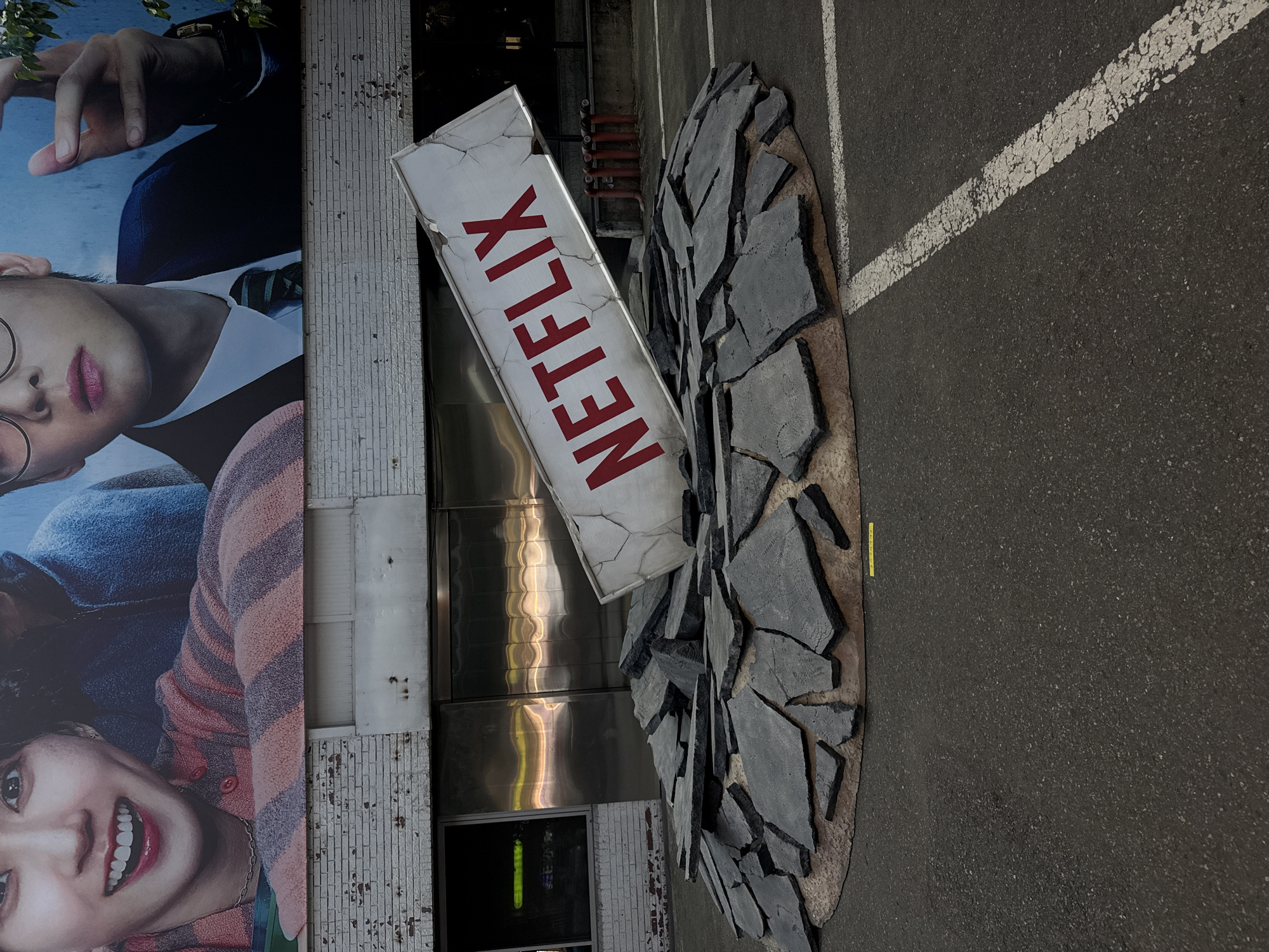 The image shows a promotional display for Netflix in Seoul, South Korea. The display features a damaged Netflix sign appearing to burst through a cracked surface on the pavement of a parking lot. The background includes a building facade with a large advertisement featuring actors. Red pipes are attached to the building behind the sign. A white painted parking line is on the right.