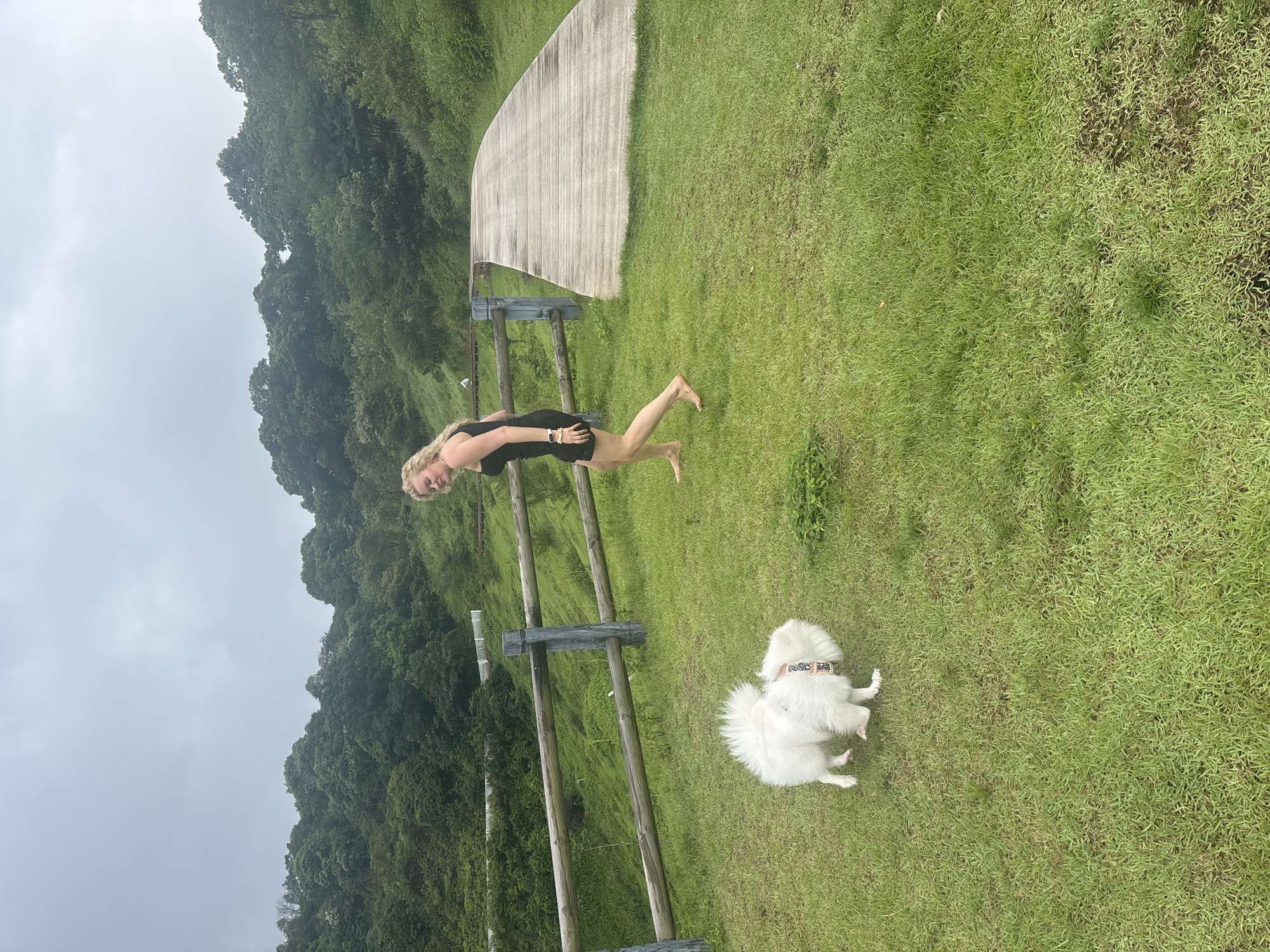 A woman with blonde hair, wearing a black sleeveless dress and barefoot, walks on a grassy area. She is looking back over her left shoulder with a smile. Next to her, to her left, a small white fluffy dog is sniffing the ground. The dog is wearing a colorful patterned collar. The scene is set outdoors with a wooden fence to the left of the woman and dog, separating them from a sloping grassy area with trees in the background. A curved wooden boardwalk extends to the right. The sky is overcast. The location is identified as Guangzhou City, China.