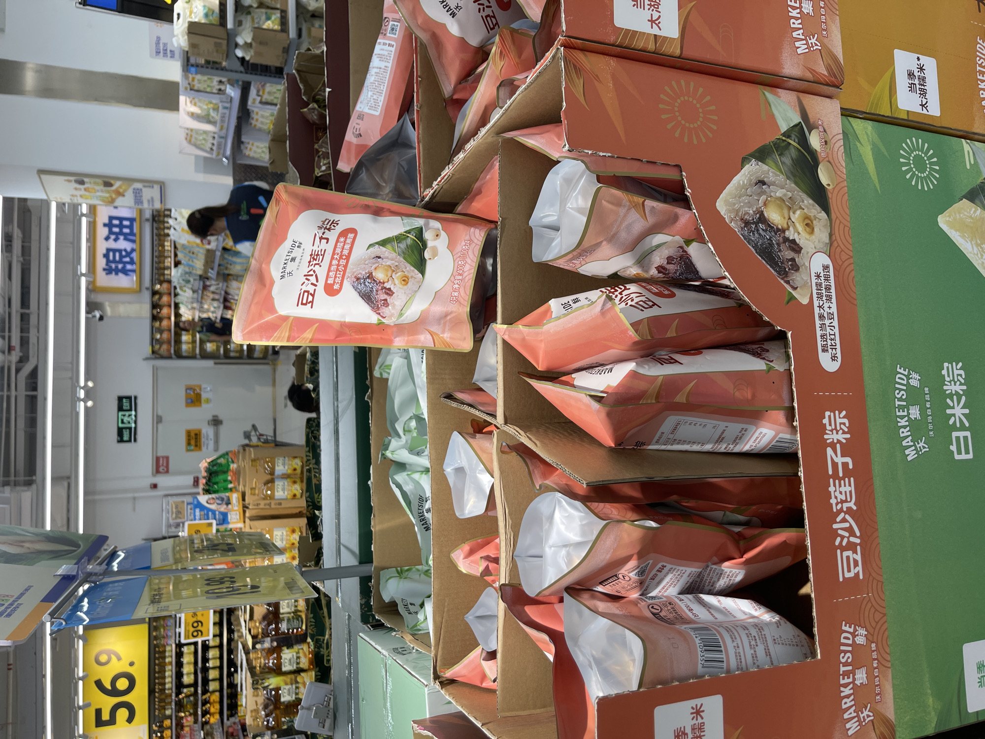 This image captures a retail display of food products, likely in a supermarket aisle, located in Panyu District, China. The foreground is dominated by cardboard boxes filled with packaged food items. One prominent package in the upper right is a pink pouch featuring a depiction of a traditional Chinese rice dumpling (zongzi) and text in Chinese characters, indicating "Soy Bean Lotus Leaf Rice Dumplings." Various other pink and white pouches are visible within the boxes, some with their tops open, revealing their contents.
In the middle ground, shelves are stocked with bottles of cooking oil and other packaged goods. Yellow signage with large black numbers "56.9" and "99.9" is visible, likely indicating prices. Behind the product display, aisles of a supermarket extend into the background, with more shelves stocked with merchandise. A woman with dark hair, wearing a blue vest, is partially visible in the background, suggesting she is a store employee. An exit sign with green illuminated characters is also visible on the wall in the background. The overall environment appears to be a well-lit, organized commercial setting.